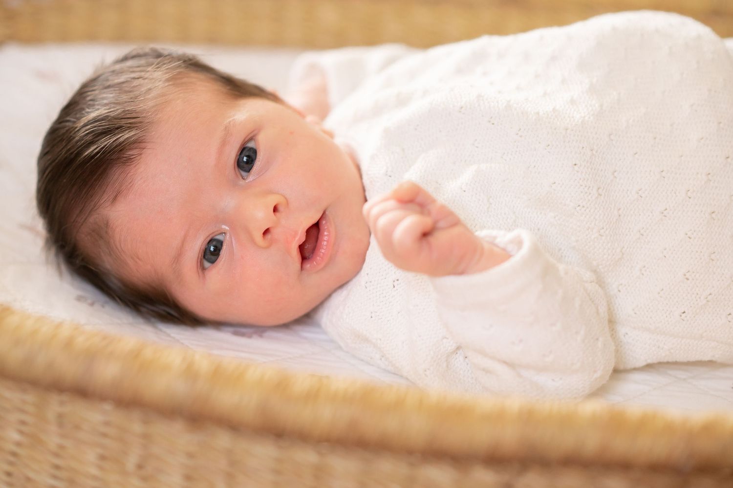 Newborn baby with dark hair lying in a wicker bassinet, wearing a white outfit, with bright blue eyes.