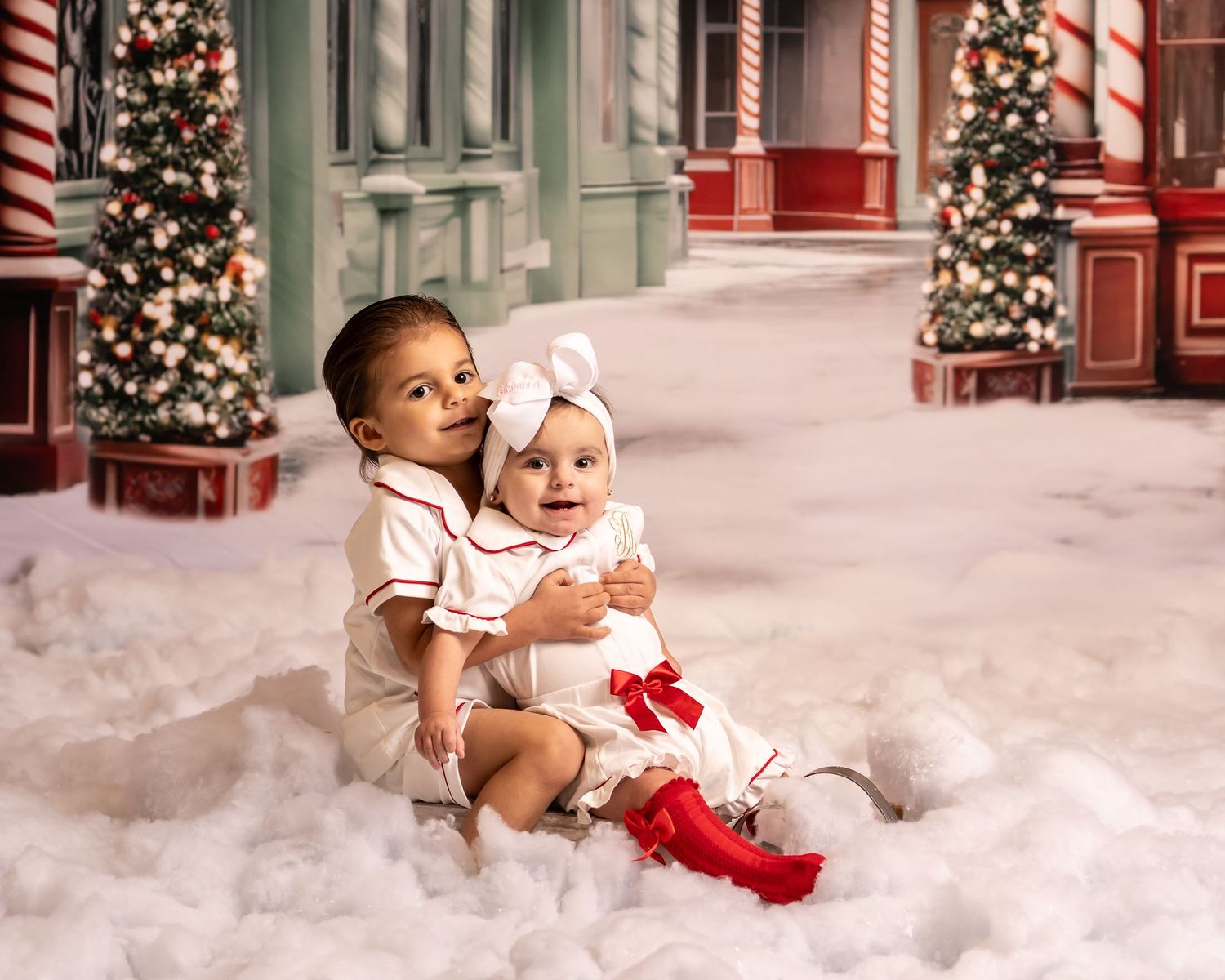 a little brother and sister in white and red pajamas sit in the fluffy snow with a Christmas scene behind them and smile for a portrait