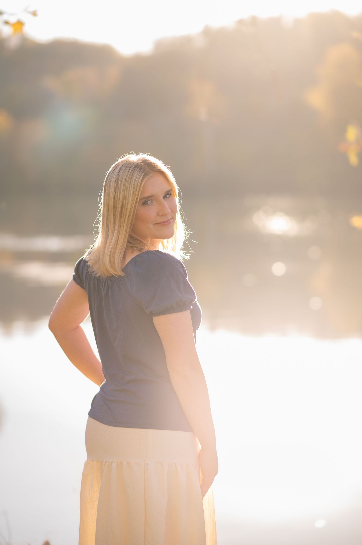 hazy, sun-drenched portrait of a blonde high school graduate standing in front of a river, looking back over her shoulder at the camera