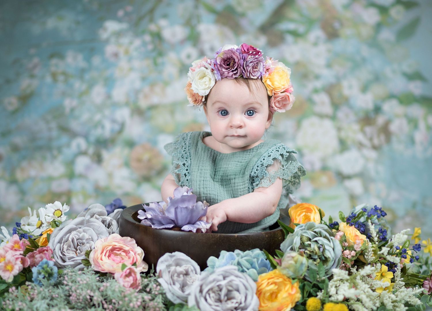 portrait of a baby girl seated in a large bowl with flowers piled around her, a flower crown, and a blue studio background