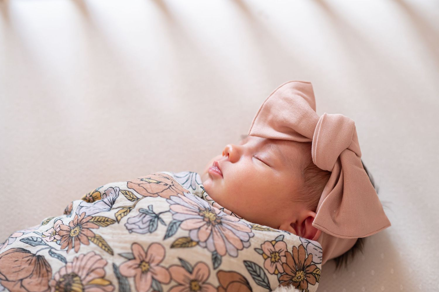 closeup newborn portrait of a baby girl swaddled in a floral wrap and pink bow, lying in her crib with light streaming through the rails