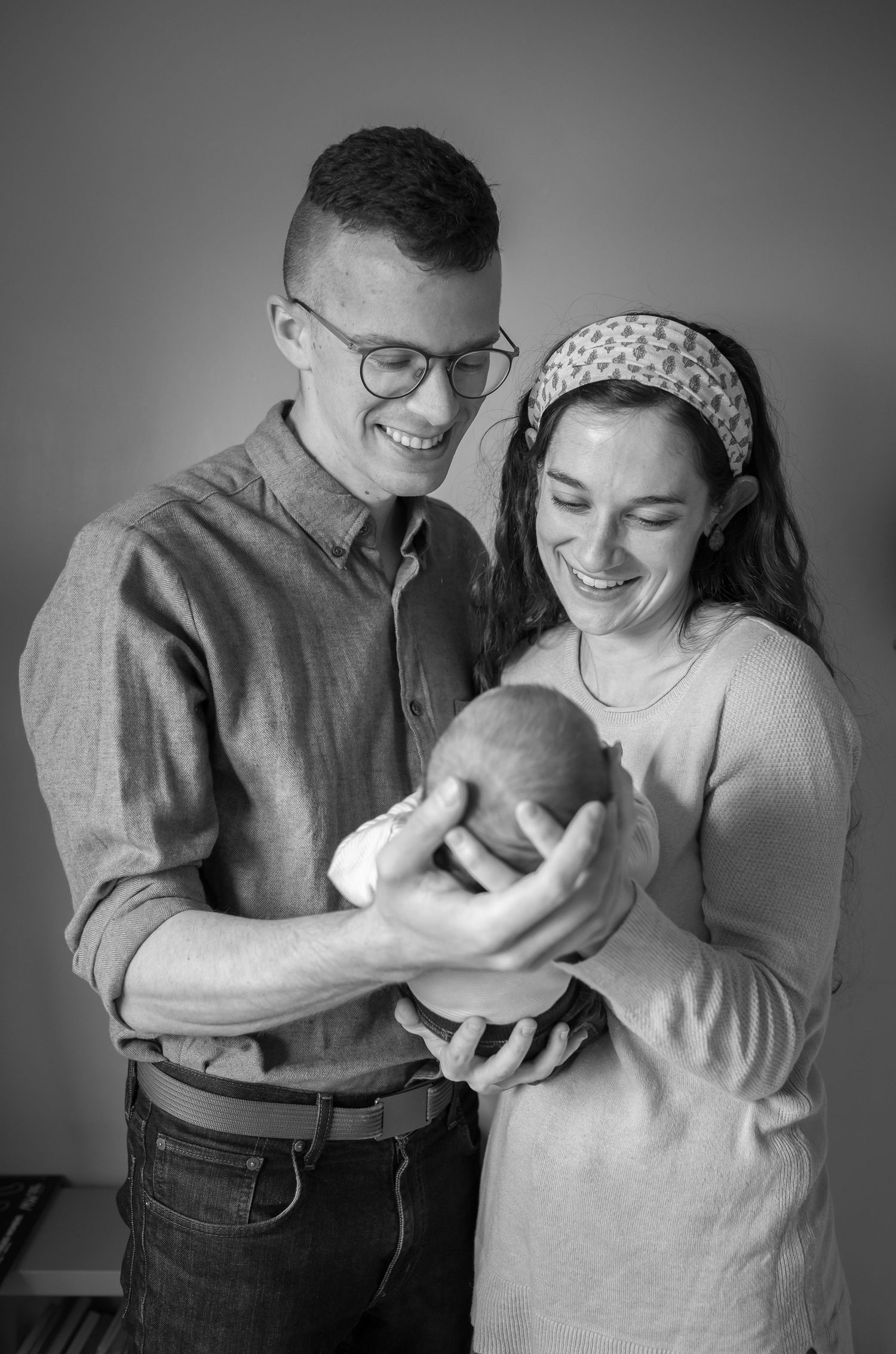 black and white portrait of a young couple standing together holding and admiring their newborn baby