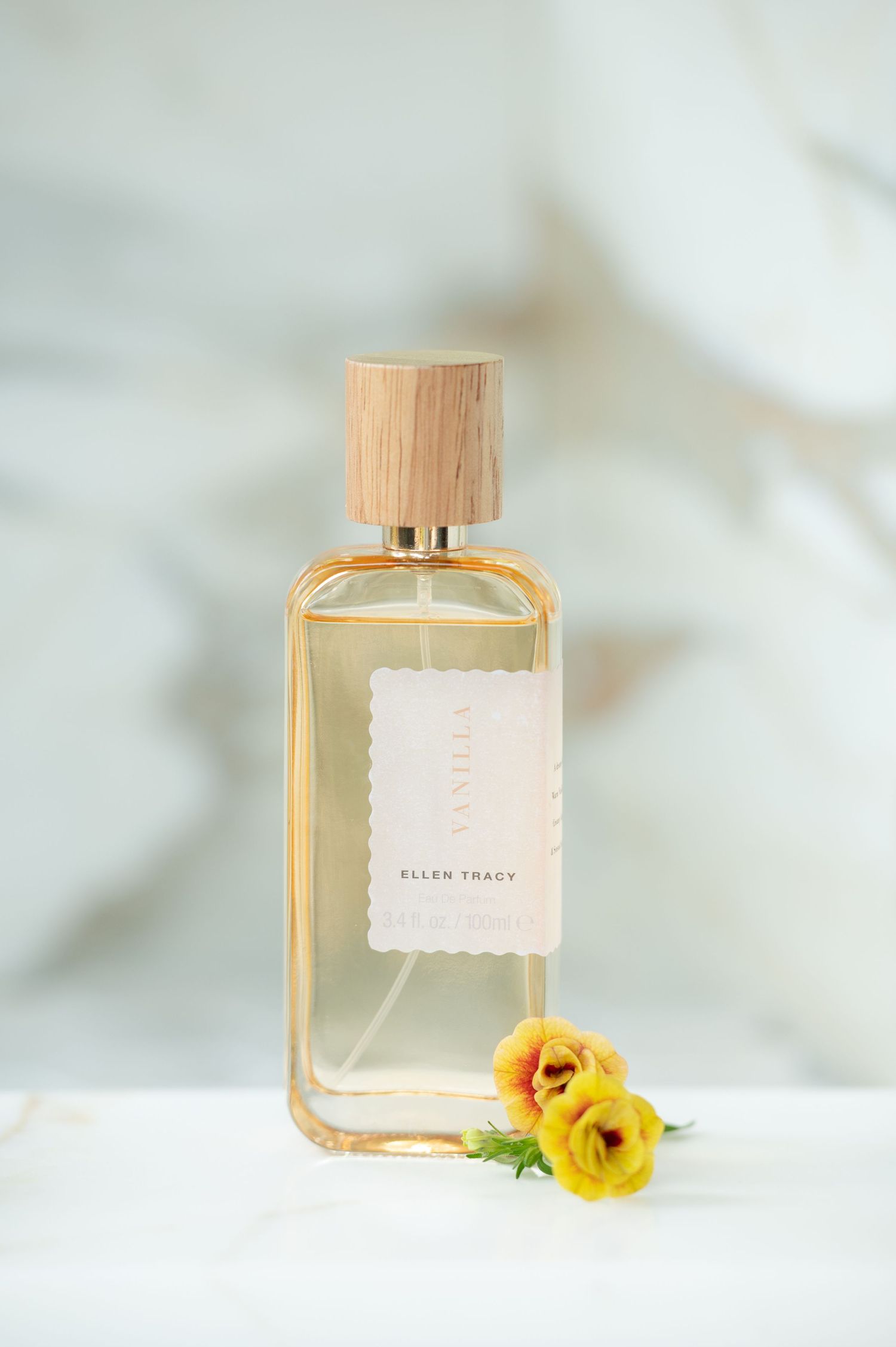 product image of a bottle of gold perfume sitting on a white marble surface with a marble wall in the background, and little yellow flowers