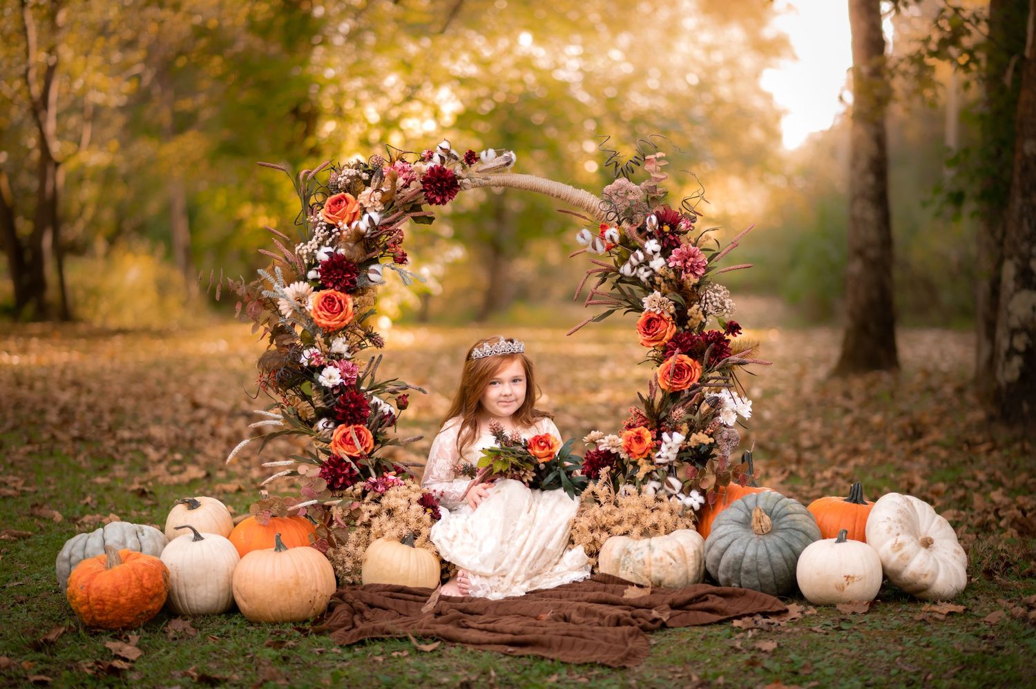 a pretty little redheaded girl in a white lace dress sits in an elaborate floral ring surrounded by pumpkin with fall colors in background
