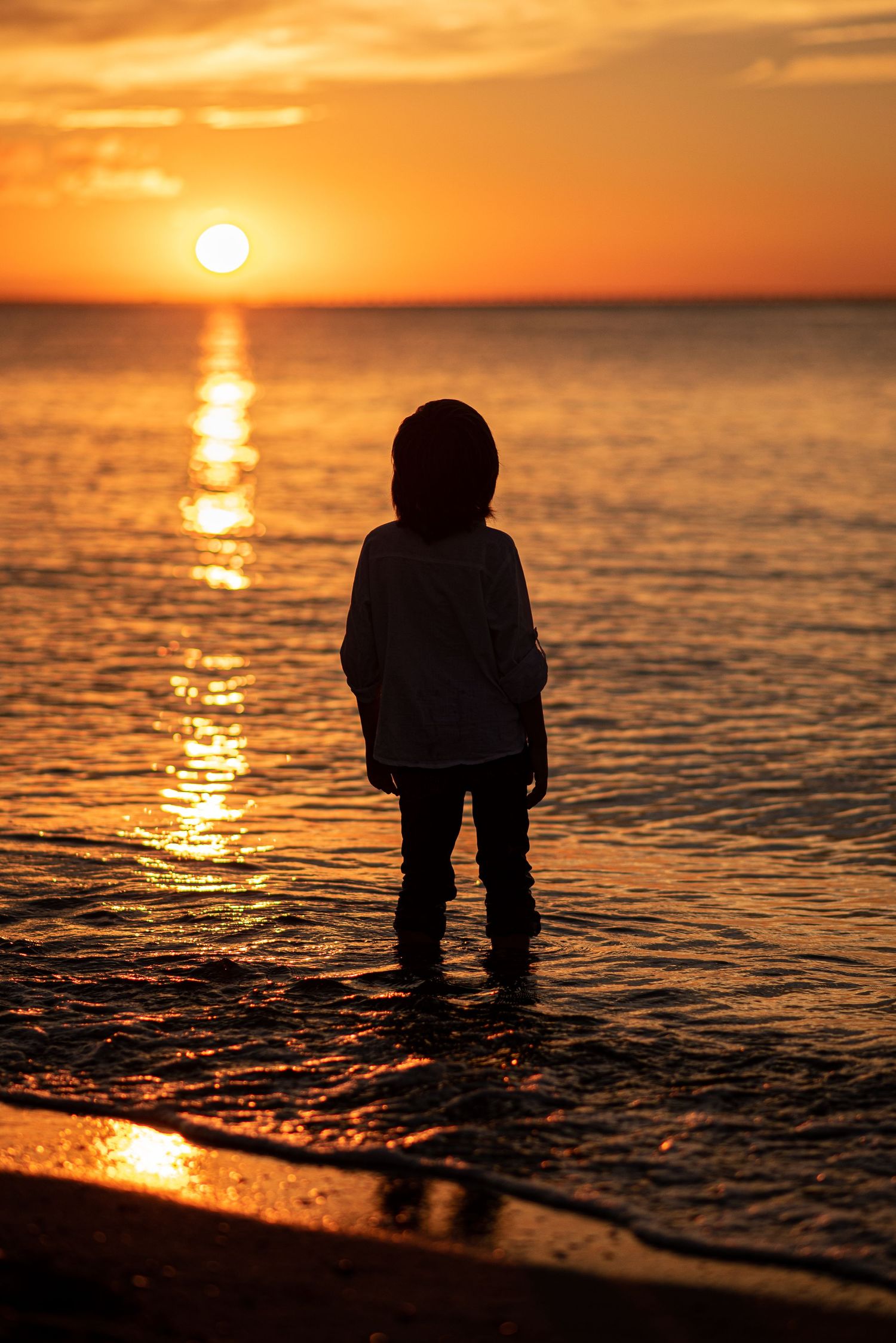 silhouette of a young boy standing on the seashore at sunset, with ocean waves lit gold by the setting sun in the background
