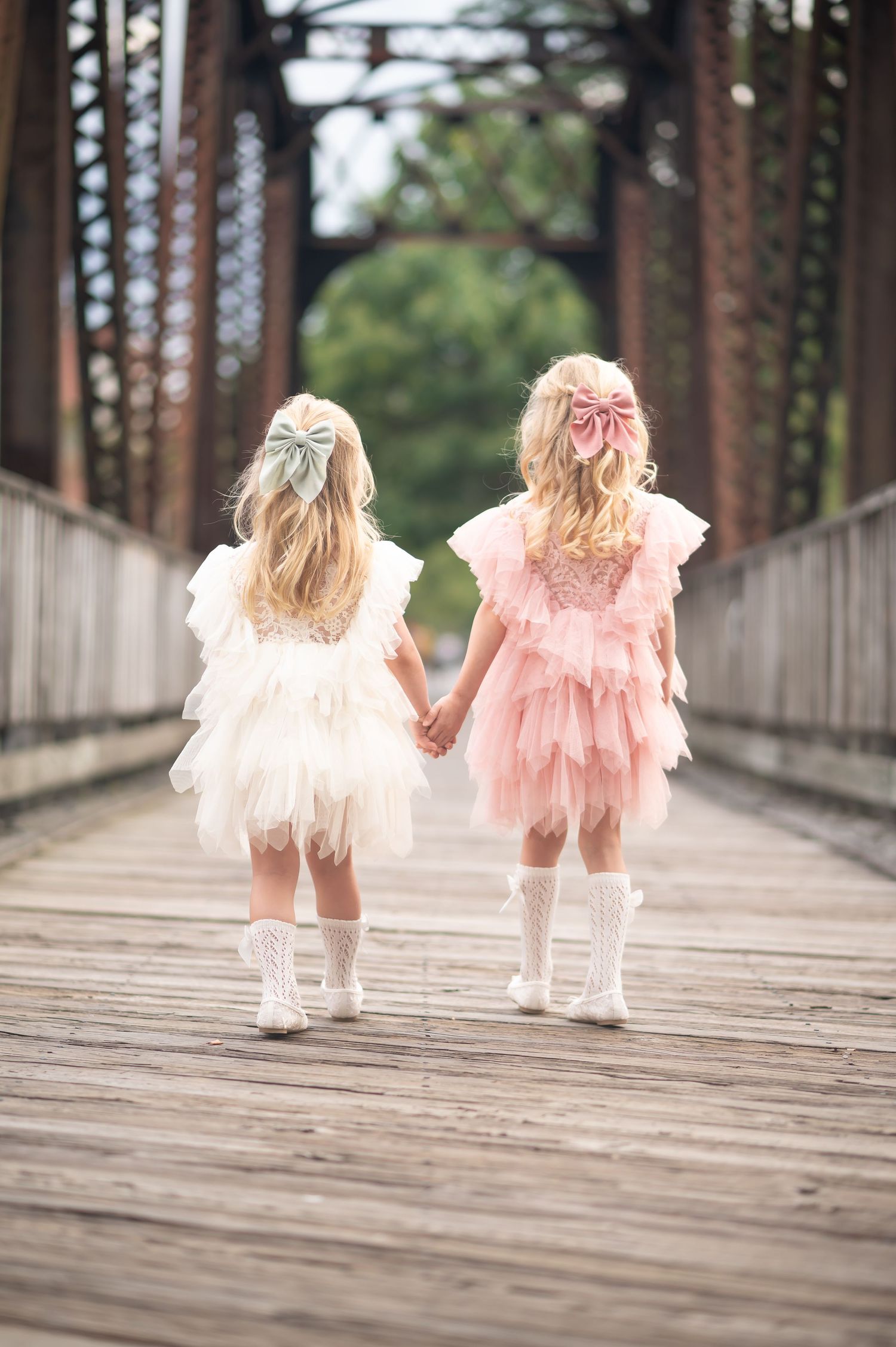 two little blonde girls in fluffy pink and white dresses walk hand in hand across an old wood and iron bridge in downtown Morgantown, wV