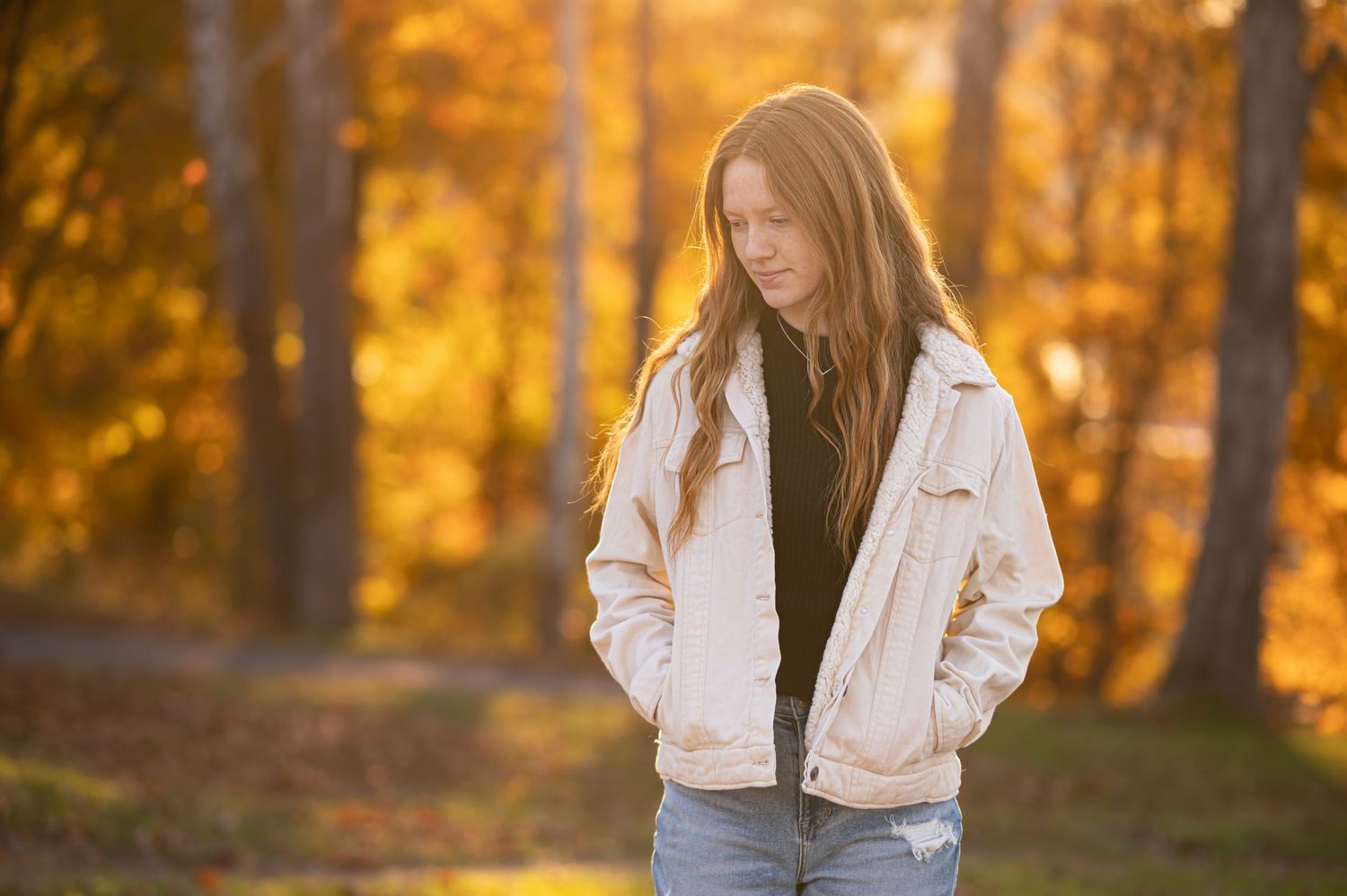 a young woman with long red hair and a white jacket strolls with her hands in her pockets and beautiful autumn foliage behind.
