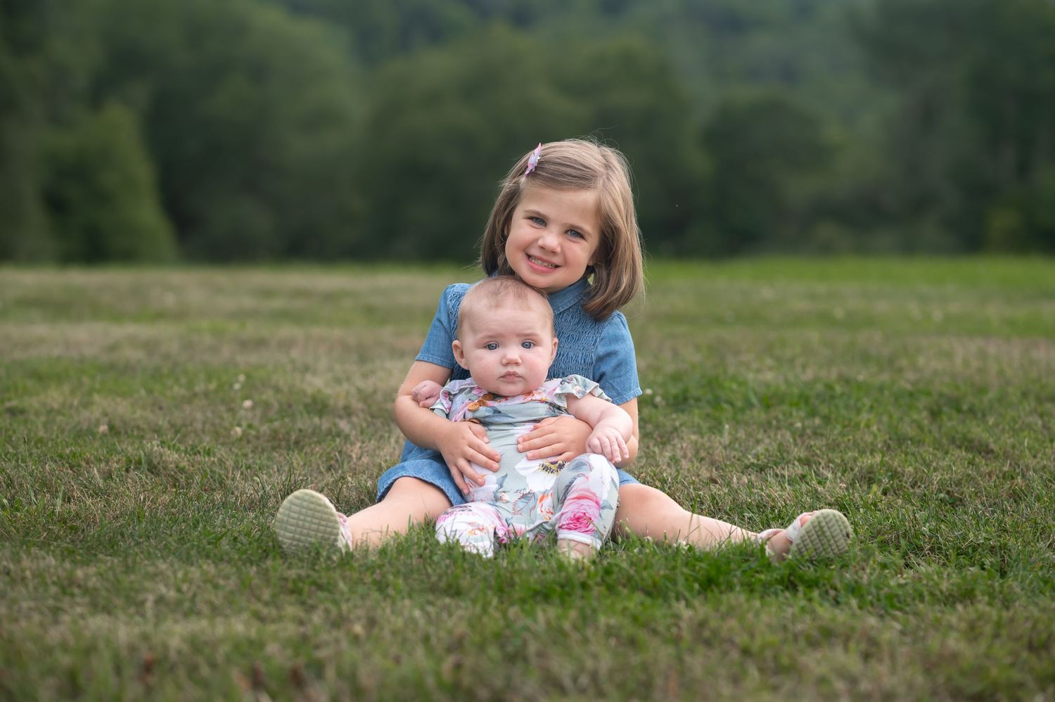 a big sister and little sister sit together in the grass and smile for a picture, with trees in the background