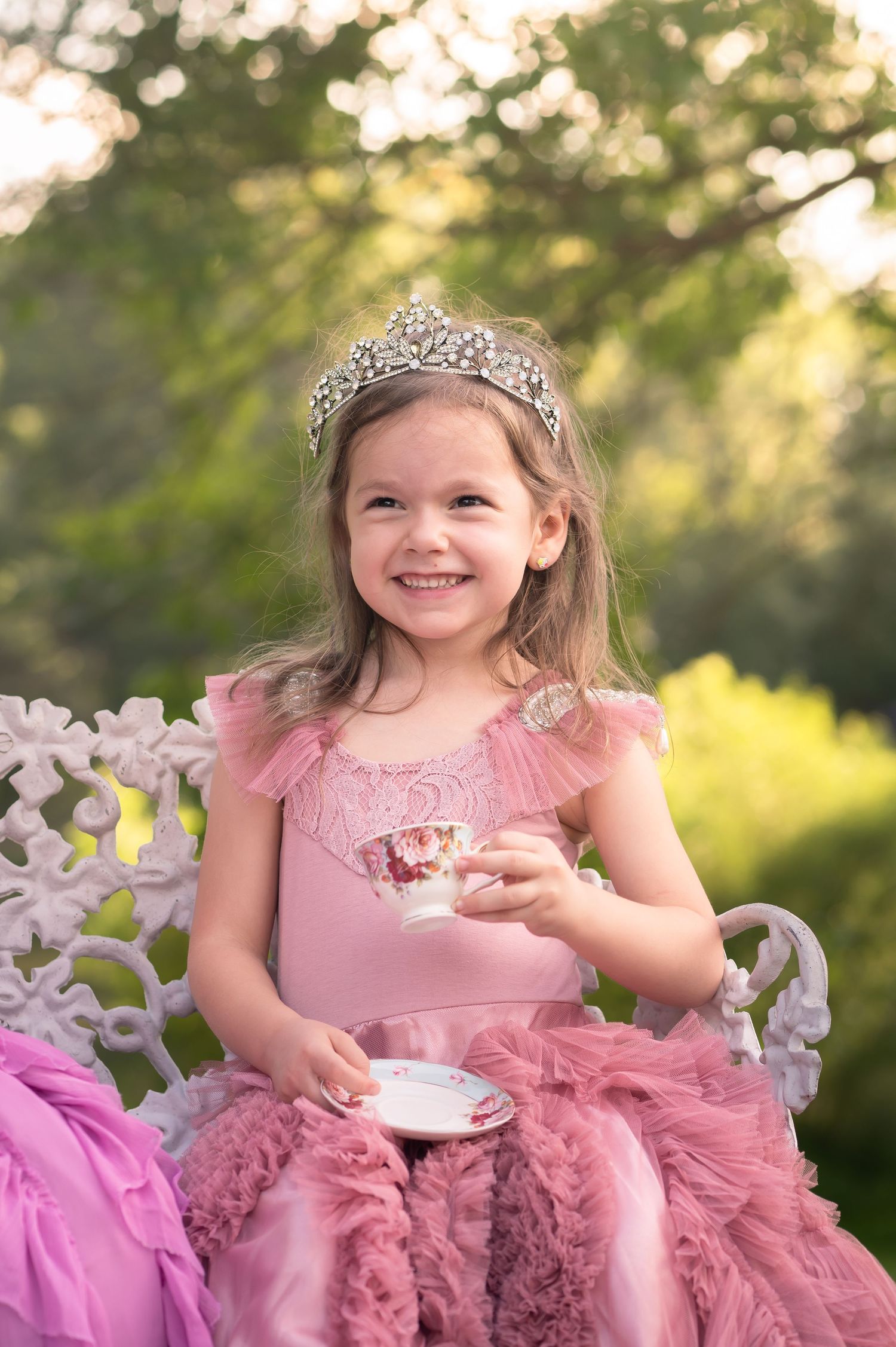 a little girl in a frilly mauve ballgown sits on a white iron bench outside and drinks tea from a tiny vintage teacup in Westover, WV