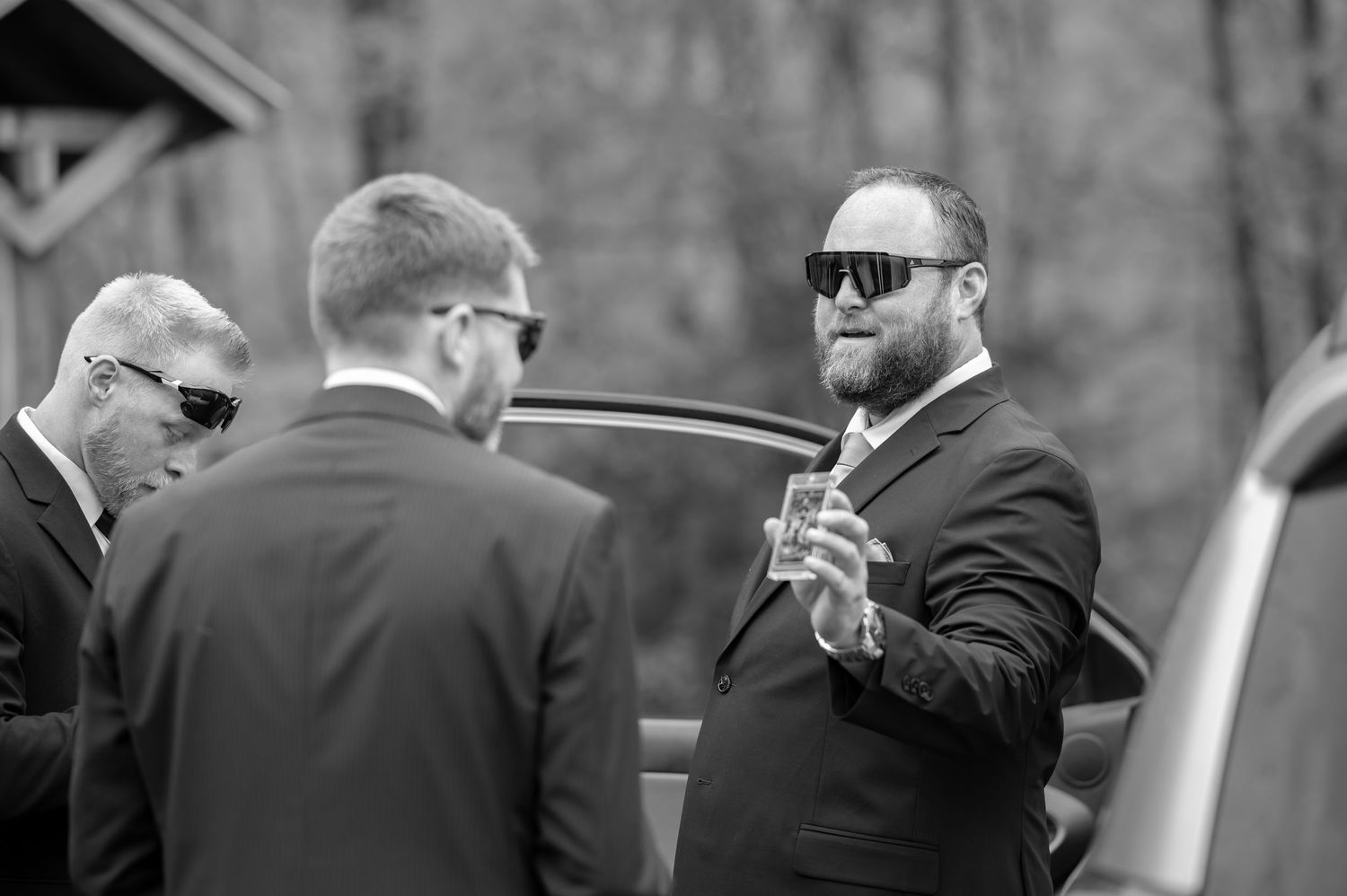 candid black and white image of a groom in sunglasses showing his groomsmen a picture on his phone, as they laugh.