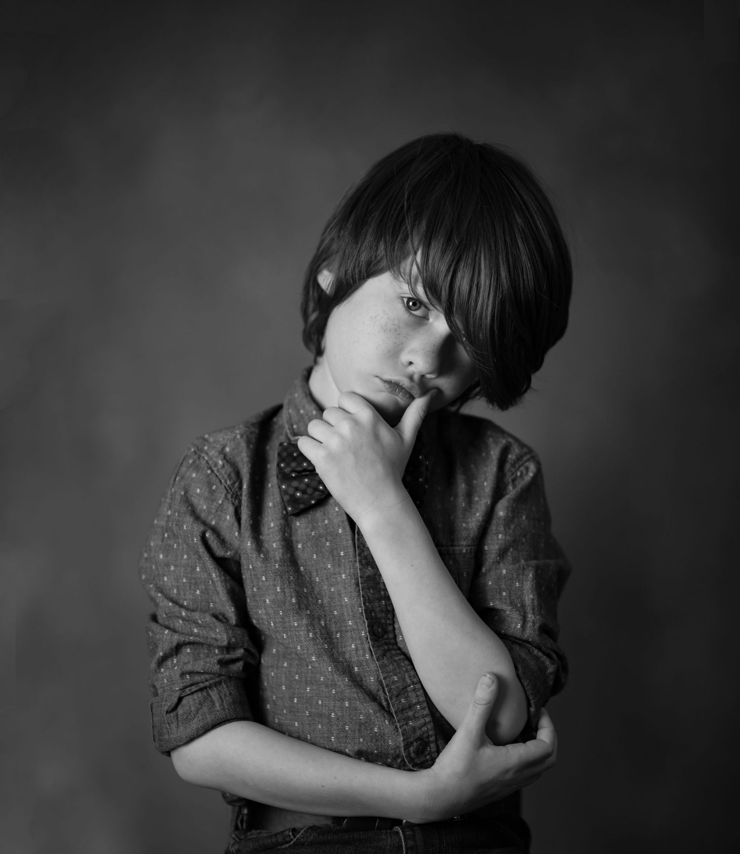black and white studio portrait of a boy with long brown hair, posing with a hand on his chin and his hair over one eye