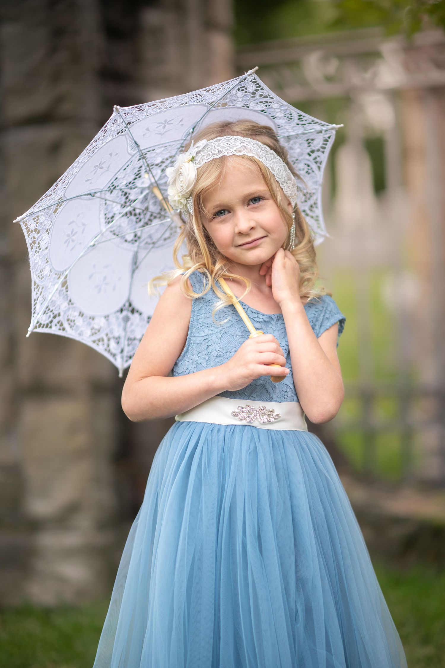 a little blonde girl in a fluffy blue dress holds a white lace parasol and smiles for a portrait