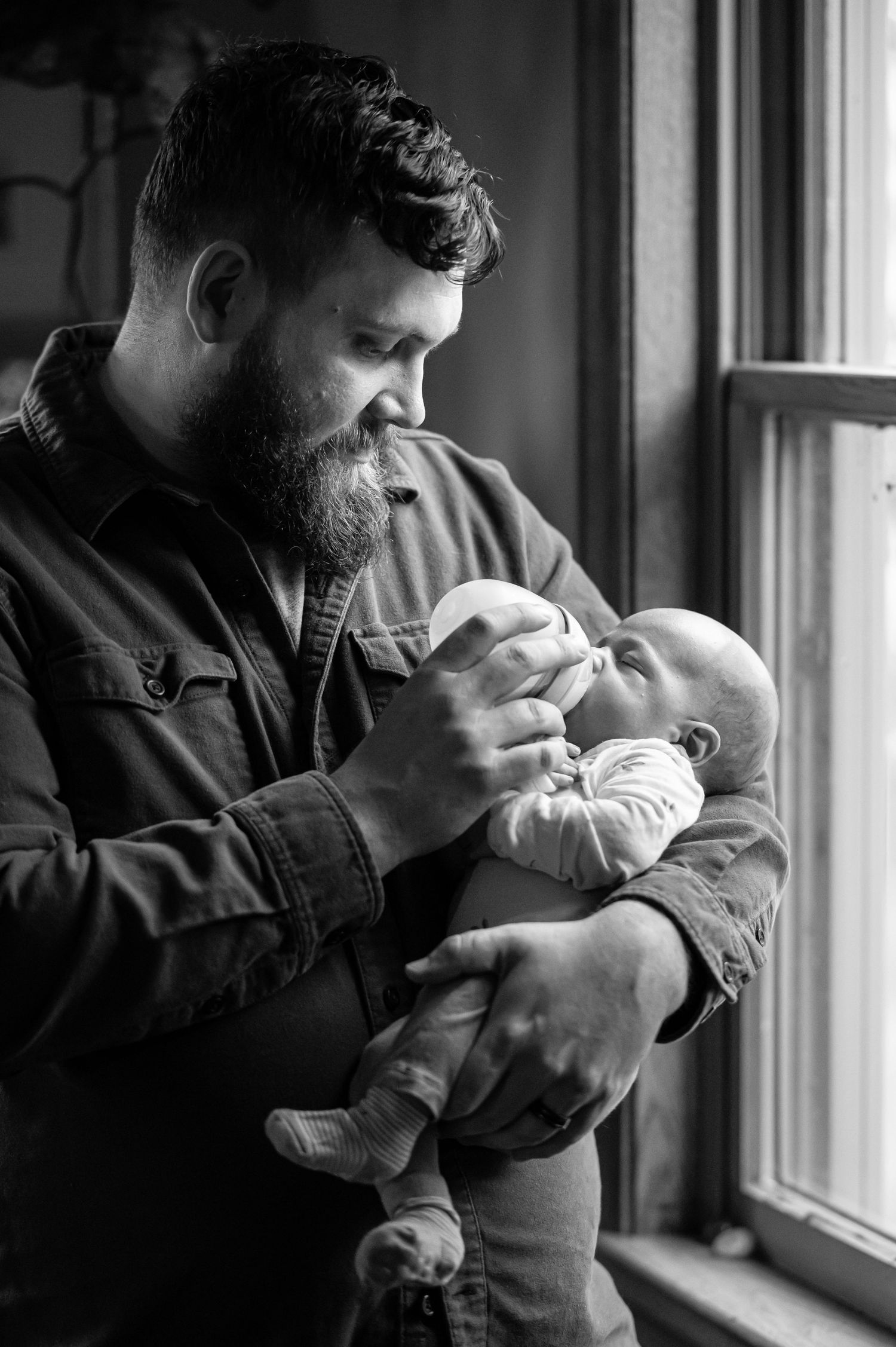 Bearded father tenderly holds and feeds newborn baby by window in black and white portrait photo.