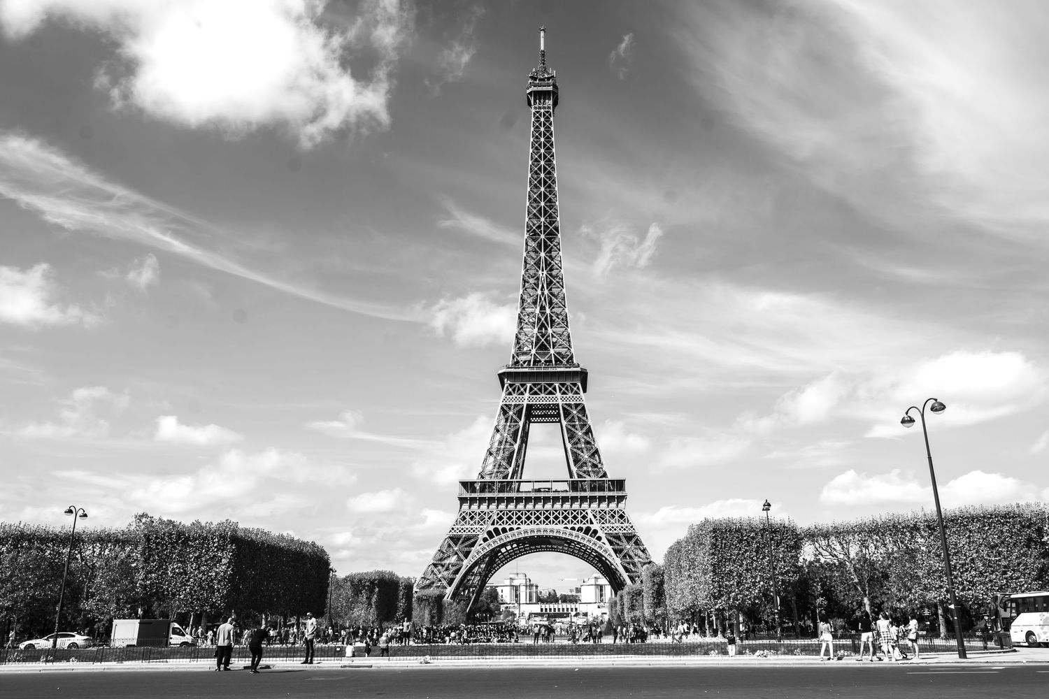 Black and white photo of the Eiffel Tower in Paris, France, with tourists and lush hedges under a cloudy sky.