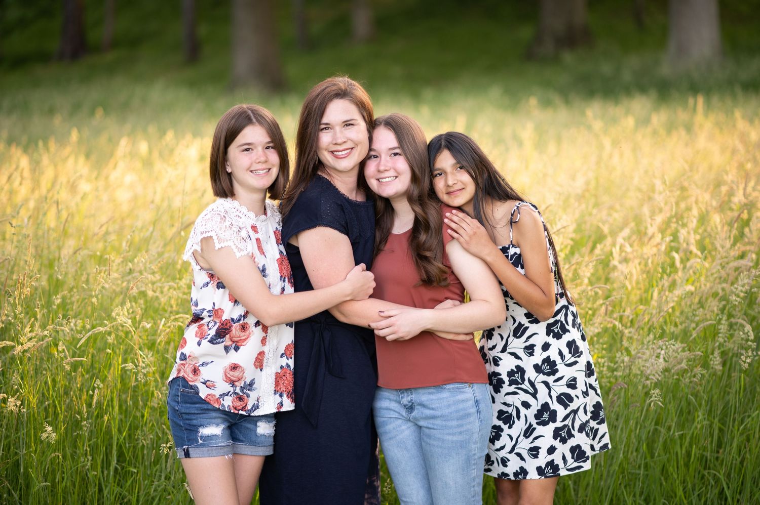 A beautiful mother and three daughters stand in field of golden grass and pose with smiles and arms around each other for a portrait