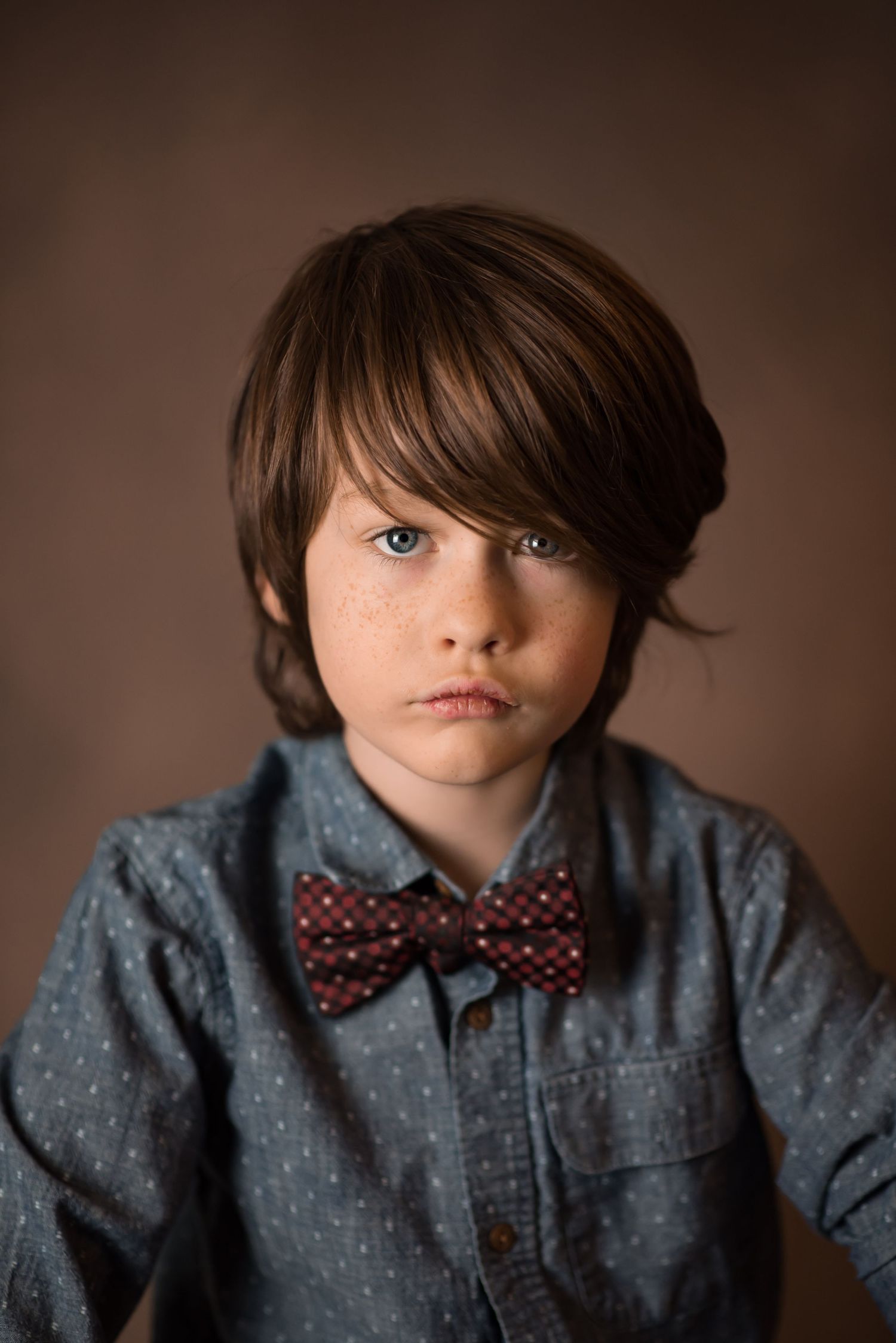 closeup headshot of a young boy in a bowtie with long brown hair over one eye, blue eyes and freckles, and a  solemn expression