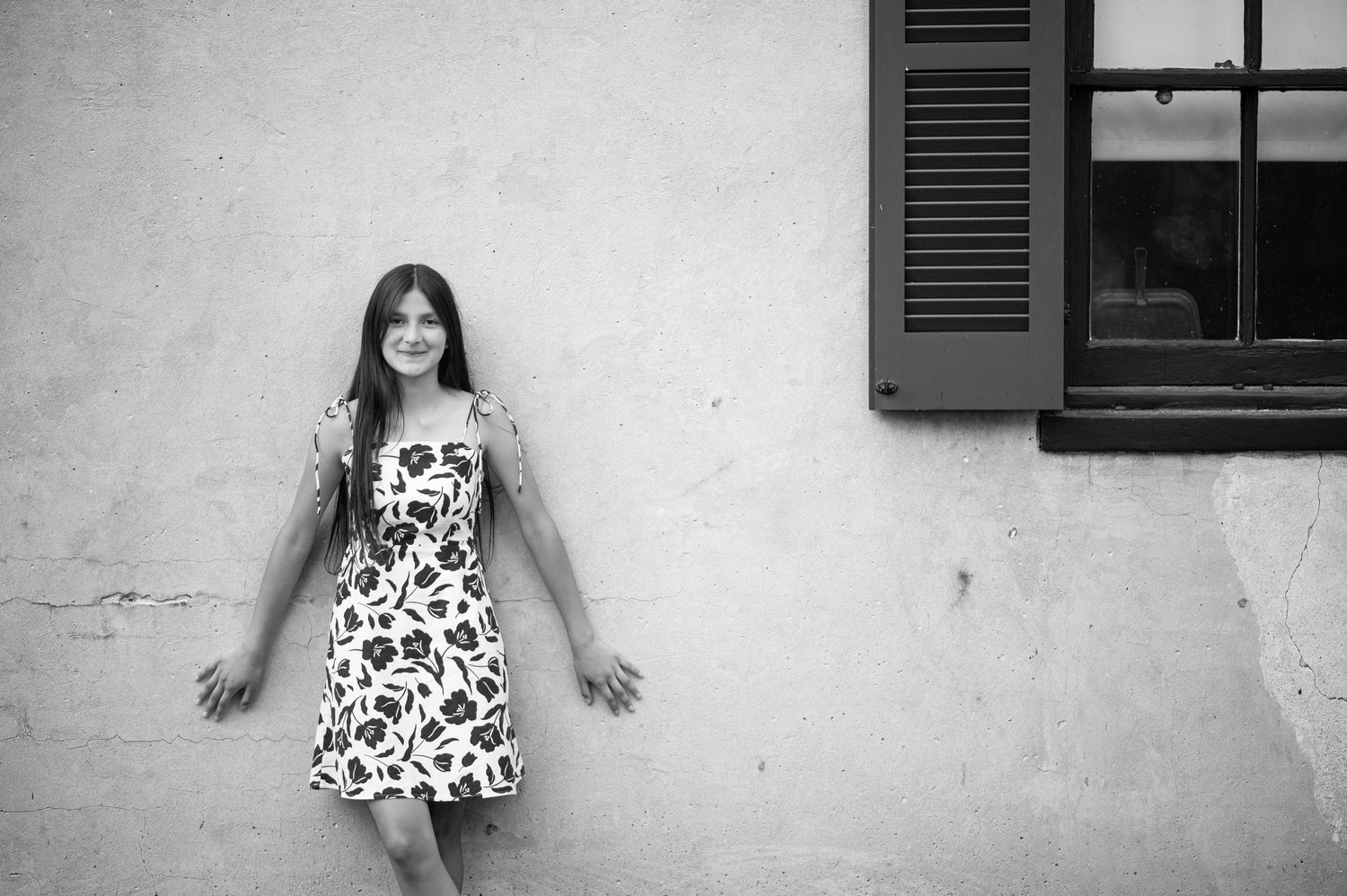 vogue black and white portrait of a young Hispanic woman in a flowered dress, standing against an old cracked wall with a window