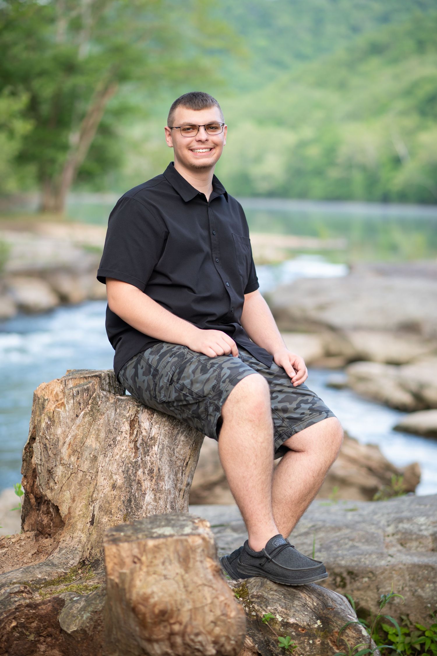 a high school graduate in a black shirt and shorts poses for a portrait on a large rock with a river and waterfall in the background