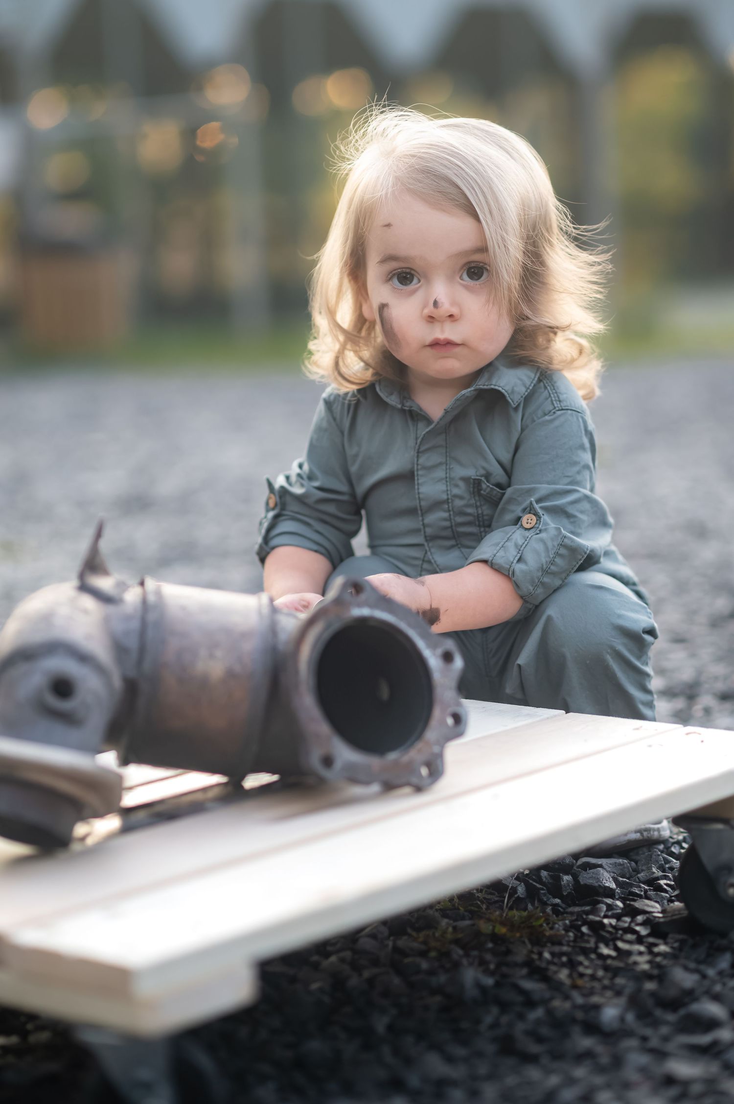 little boy with golden hair in a mechanic's uniform sits on a gravel lot holding a large piece of exhaust pipe, with grease on his nose