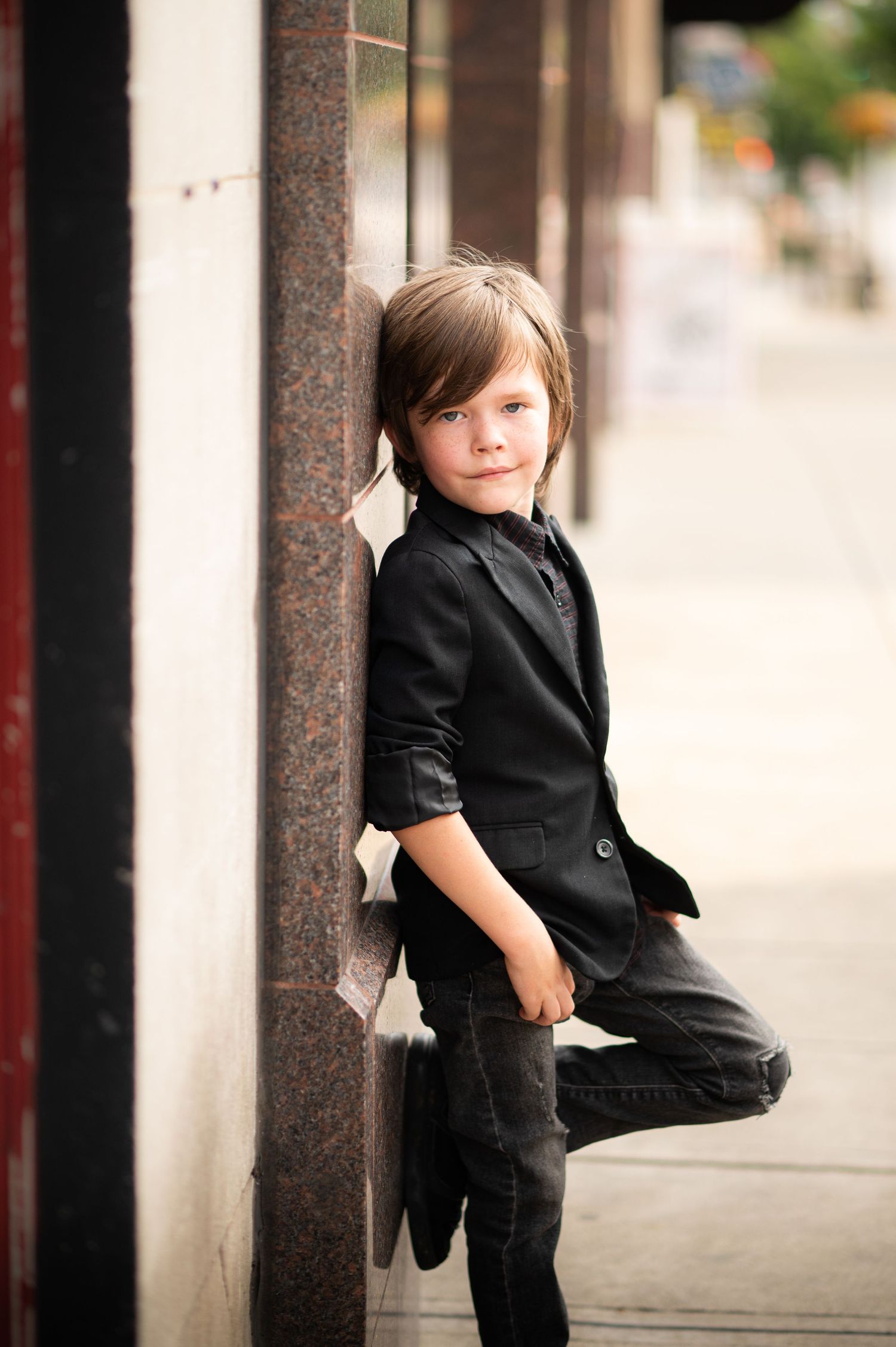 urban portrait of a little boy in a black suit, leaning up against the wall of a building in downtown Morgantown, WV and smiling