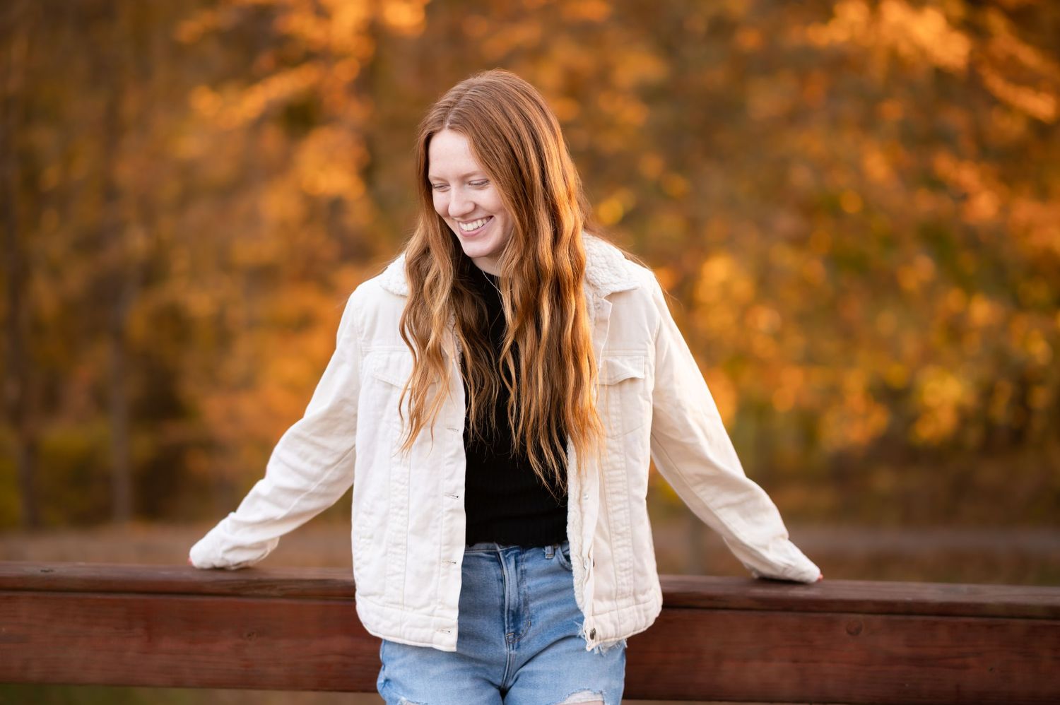 a red haired senior student in a white jacket leans back against a wooden railing and laughs, with autumn leaves in the background