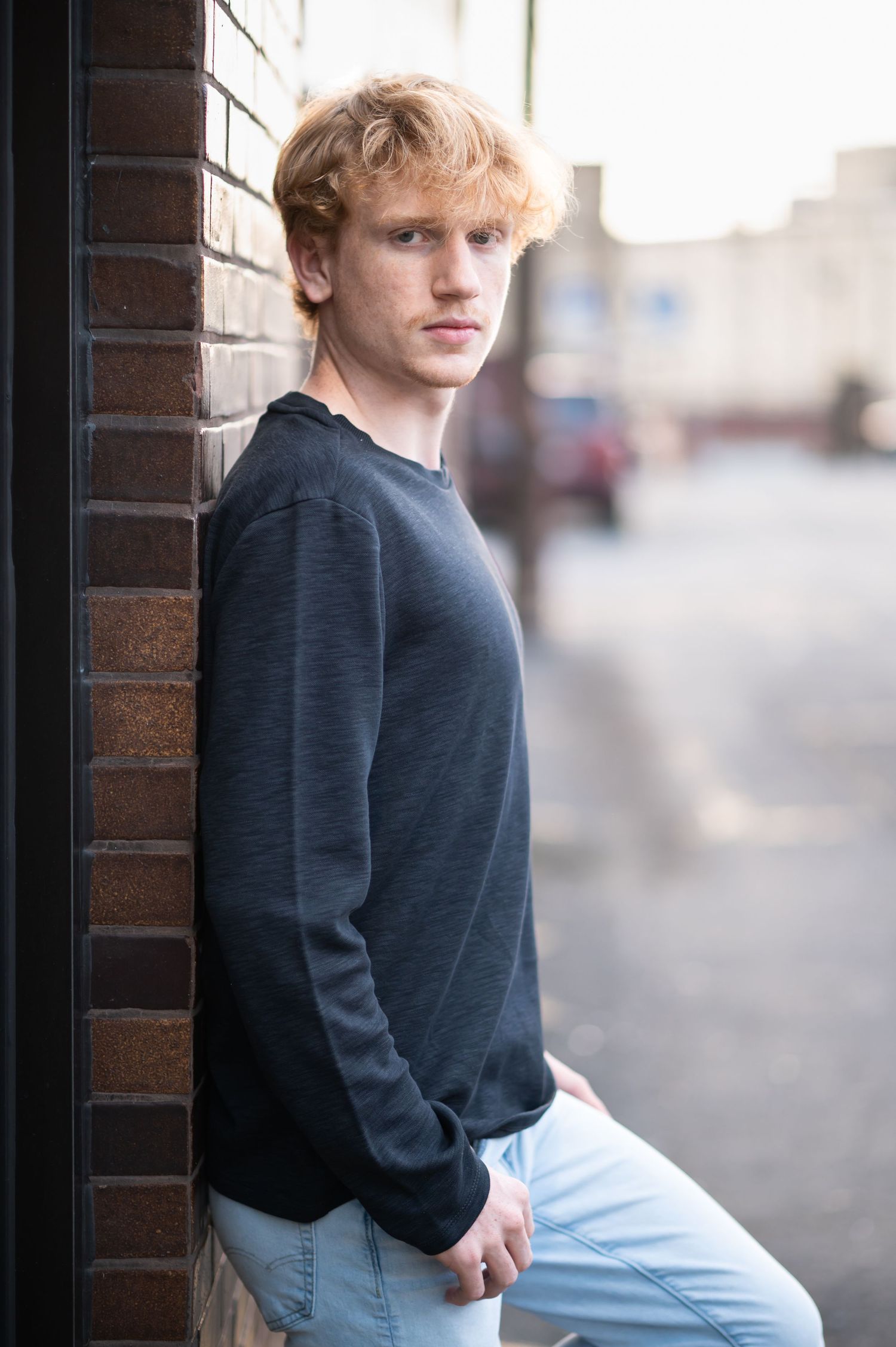 a high school student in a black top leans against a brick wall in an alley in downtown Morgantown, WV and looks solemnly at the camera