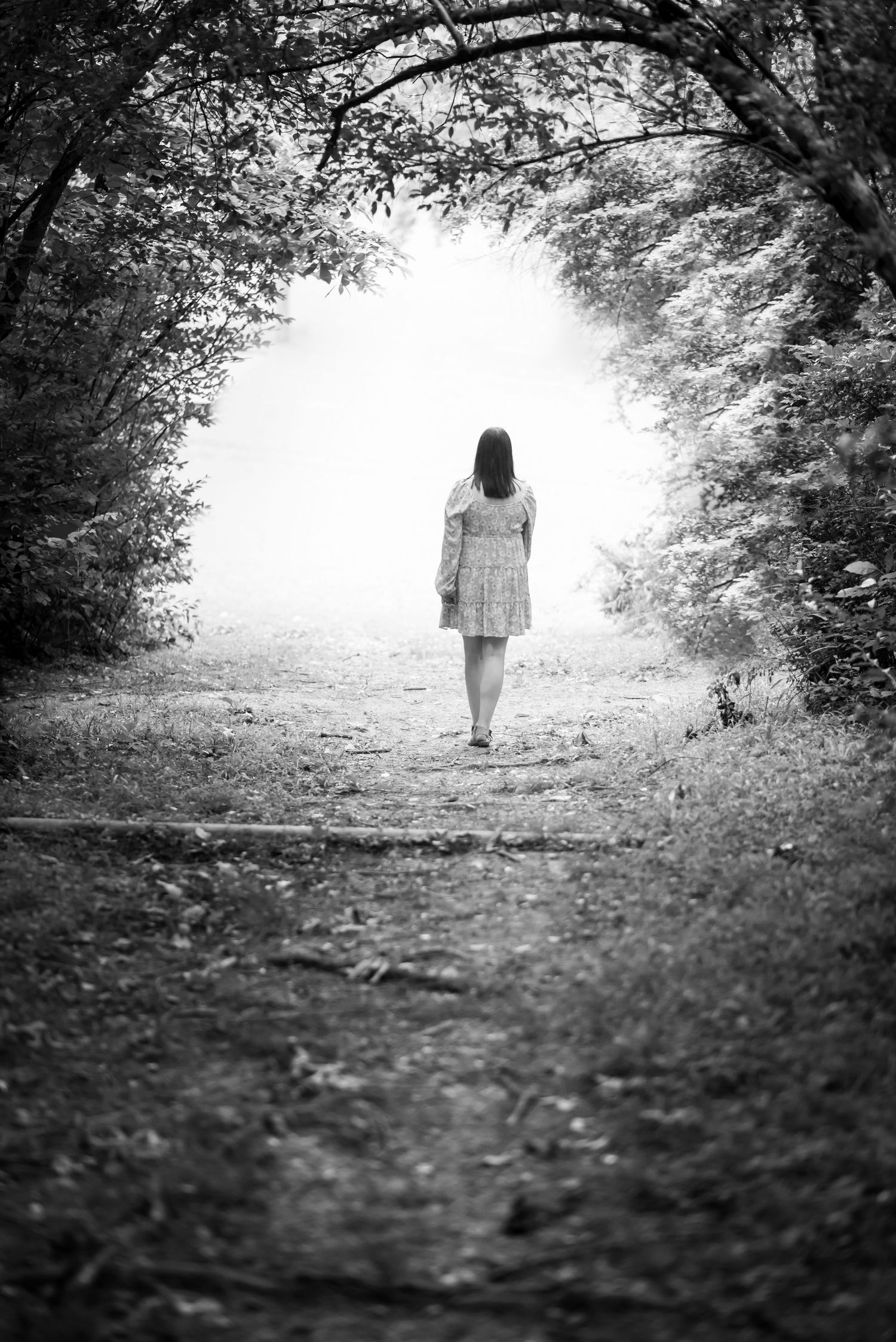 environmental black and white portrait of a young woman walking away in the distance under an arch of trees, with light in the distance
