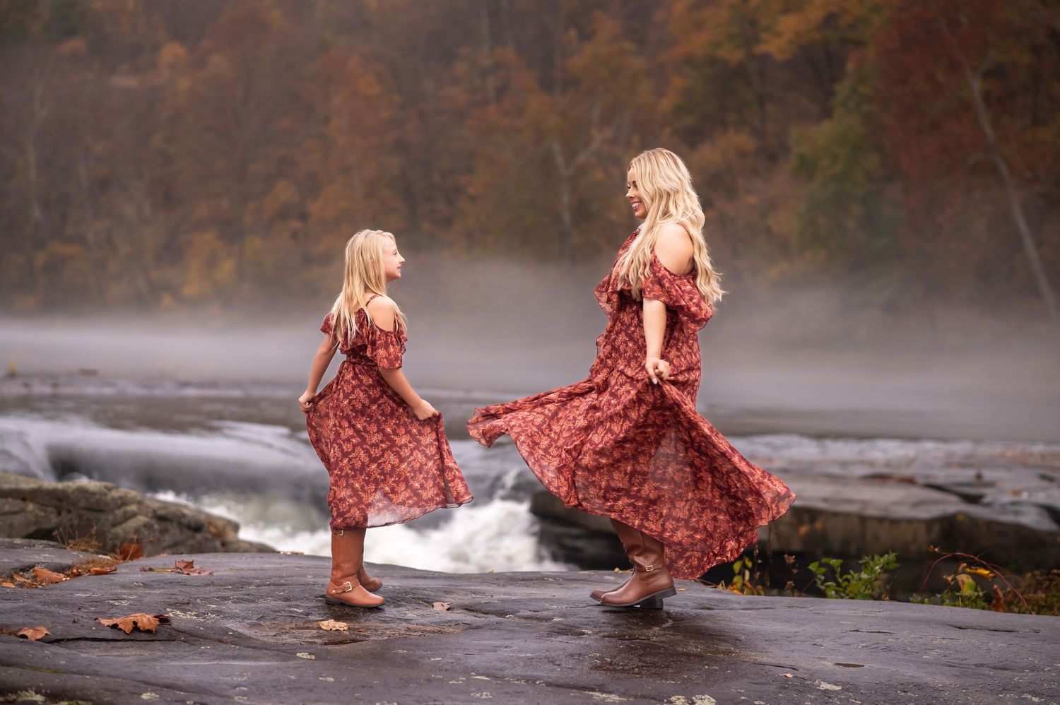 a blonde mother and daughter in matching red dresses twirl in front of a waterfall at Valley Falls, WV for fall portraits together