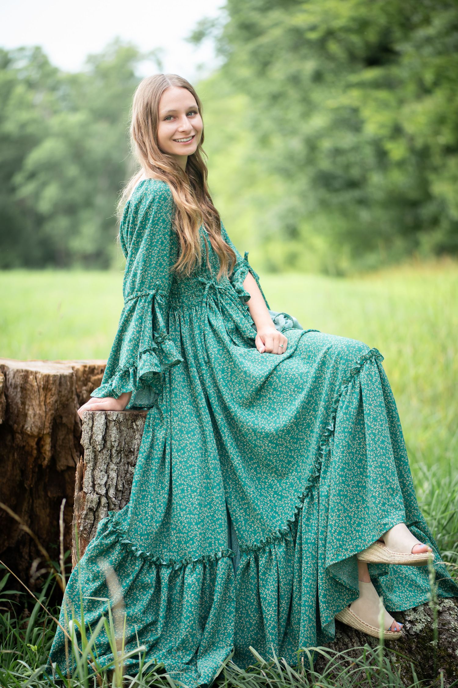 a senior student in a long green dress poses on a large tree stump for a portrait, with green fields and trees in the background
