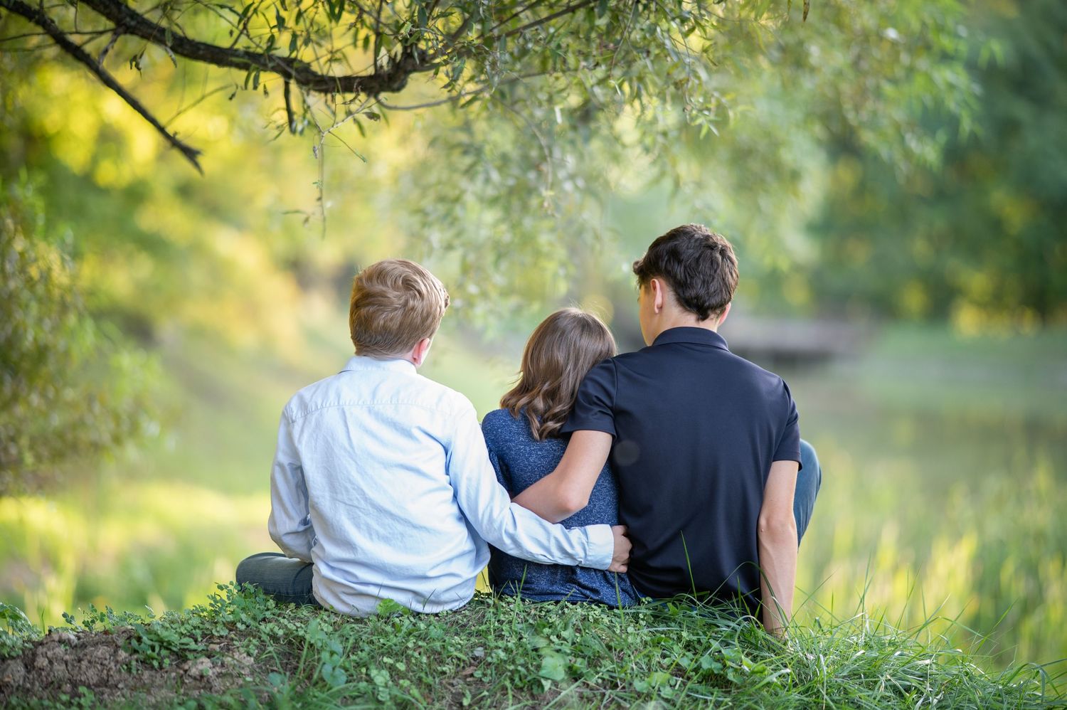 three siblings in shades of blue sit on a little hill overlooking a lake, with softly blurred, sunlit foliage in the background