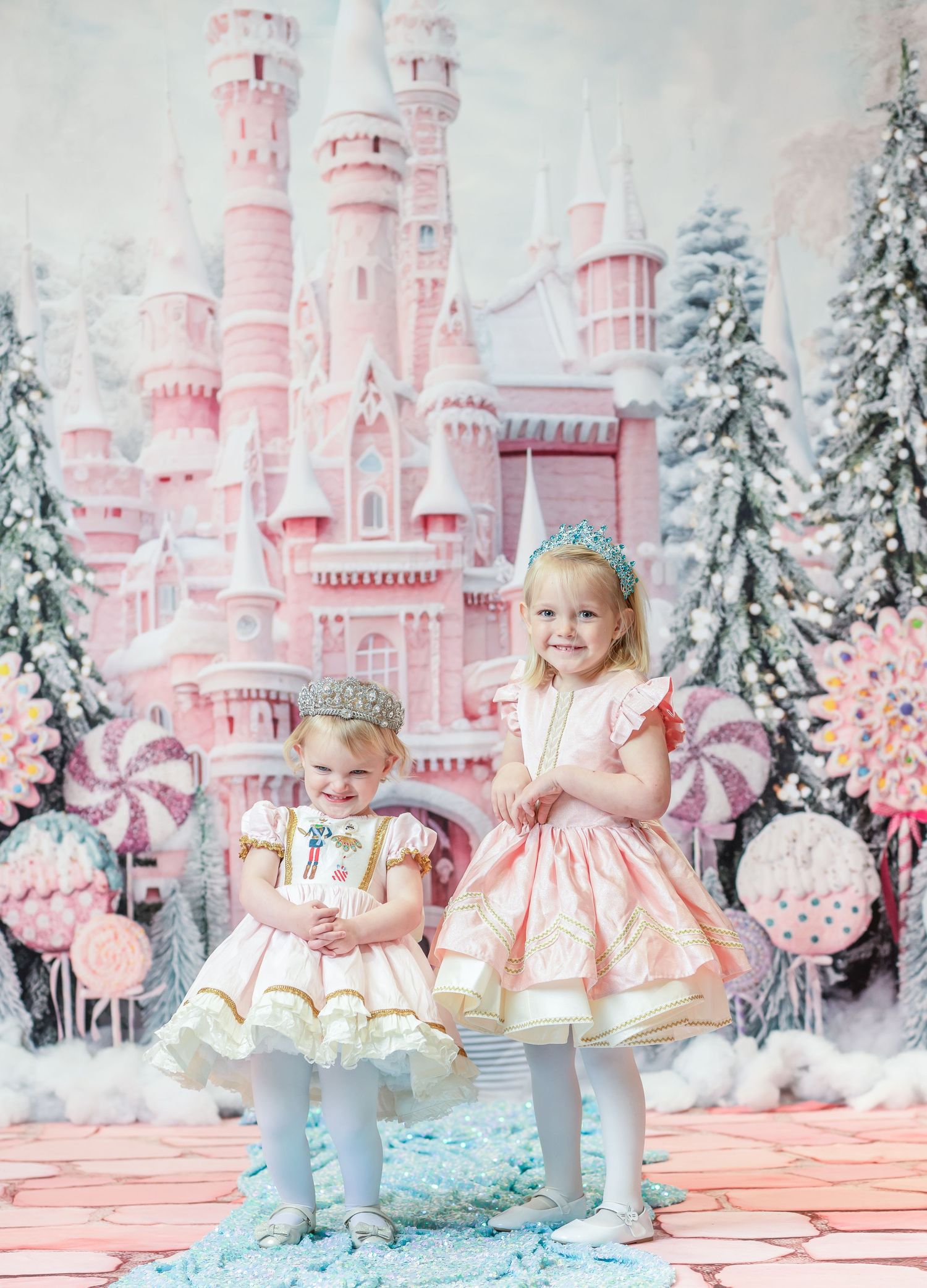 two little sisters in pink dresses stand in the snow and giggle together with a pink candy castle in the background