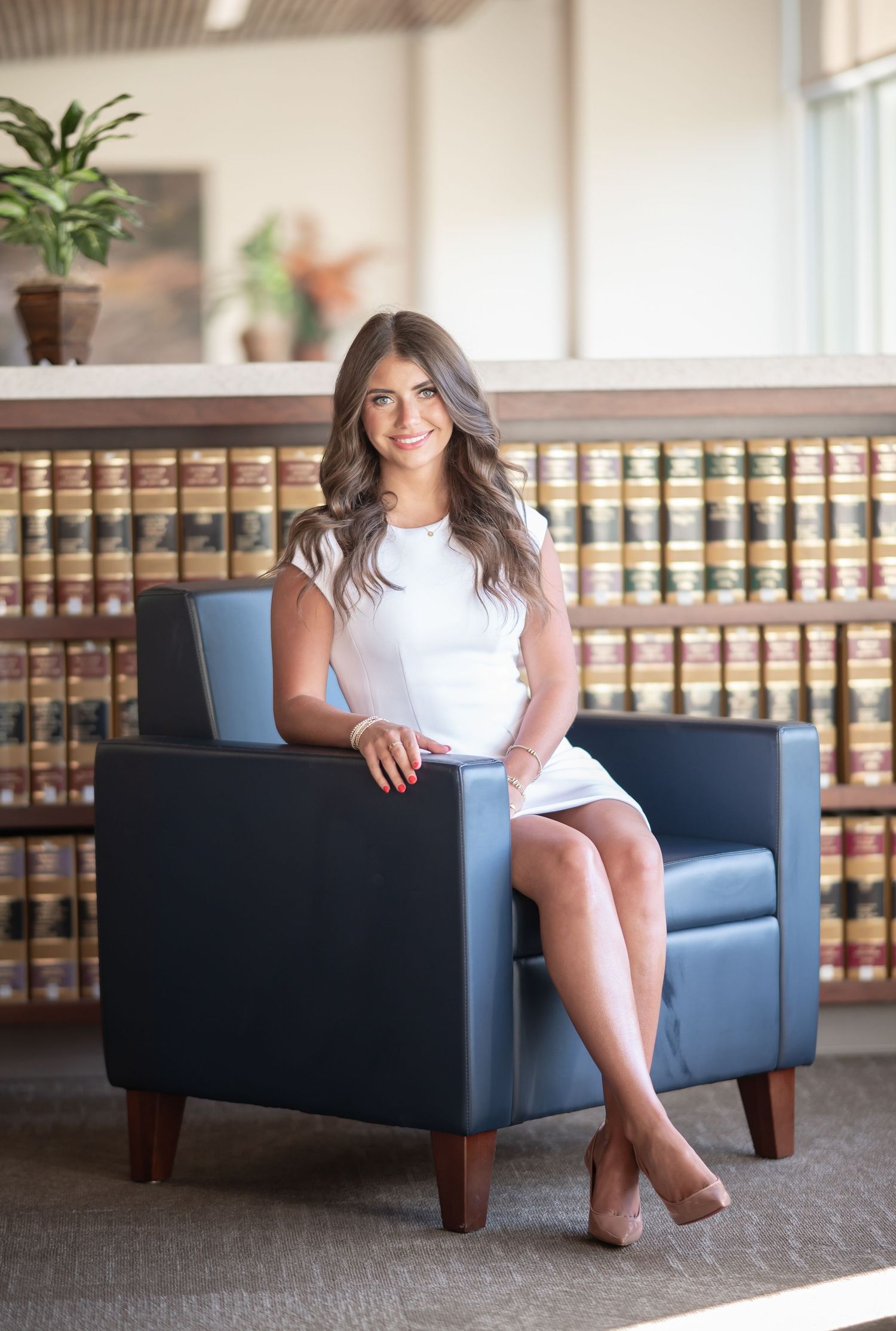college senior portrait of a graduating law student in a white dress, seated in a blue chair with shelves of law books behind her