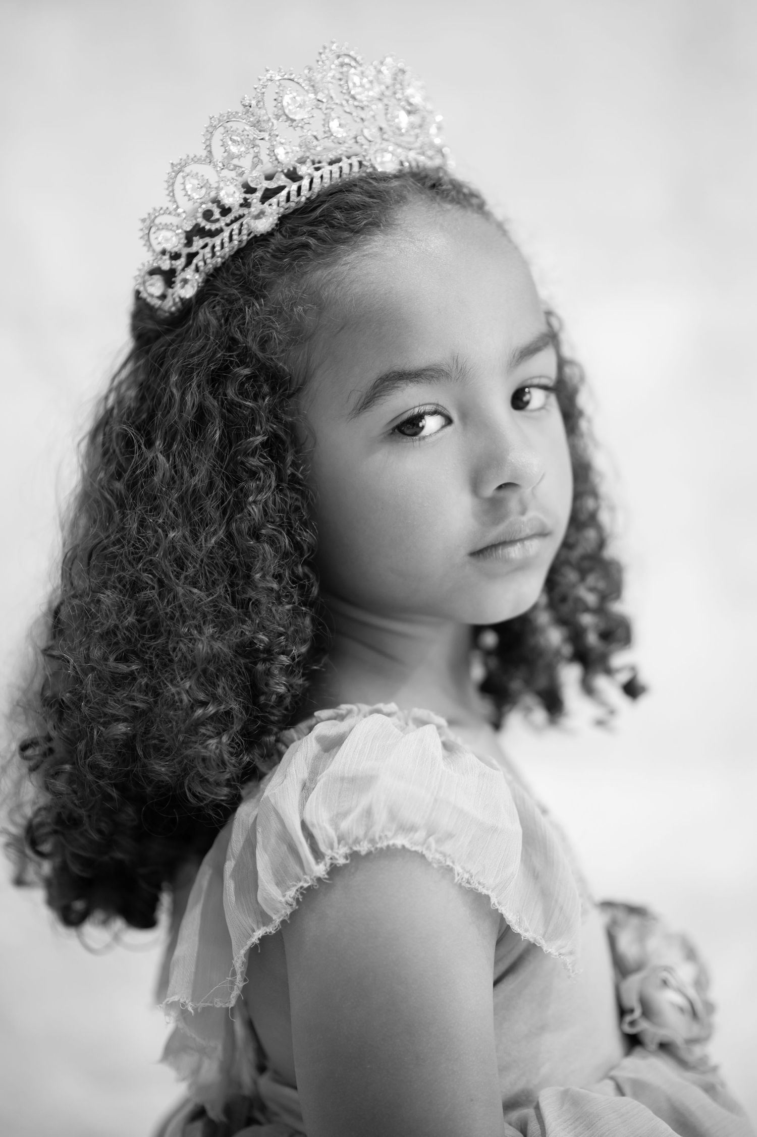 black and white closeup portrait of a little girl in a dress and crown, looking over her shoulder at the camera with a serious expression