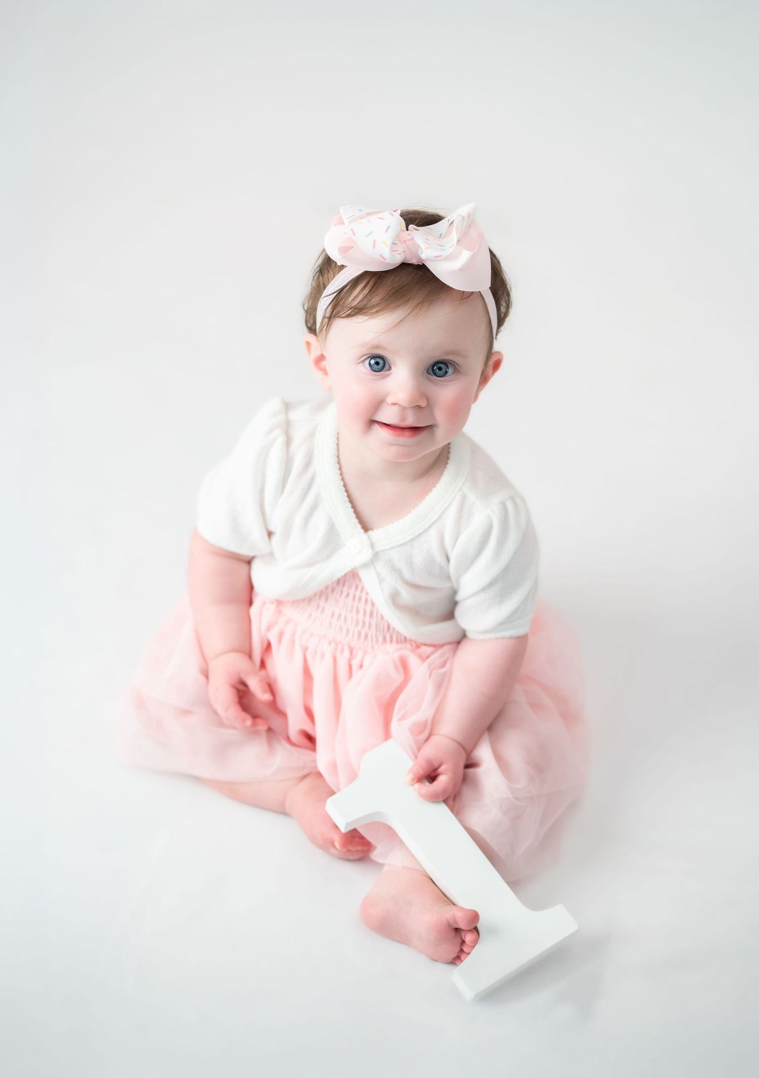 a little girl in a pink and white dress sits on a white studio background and smiles while holding a white wooden number one