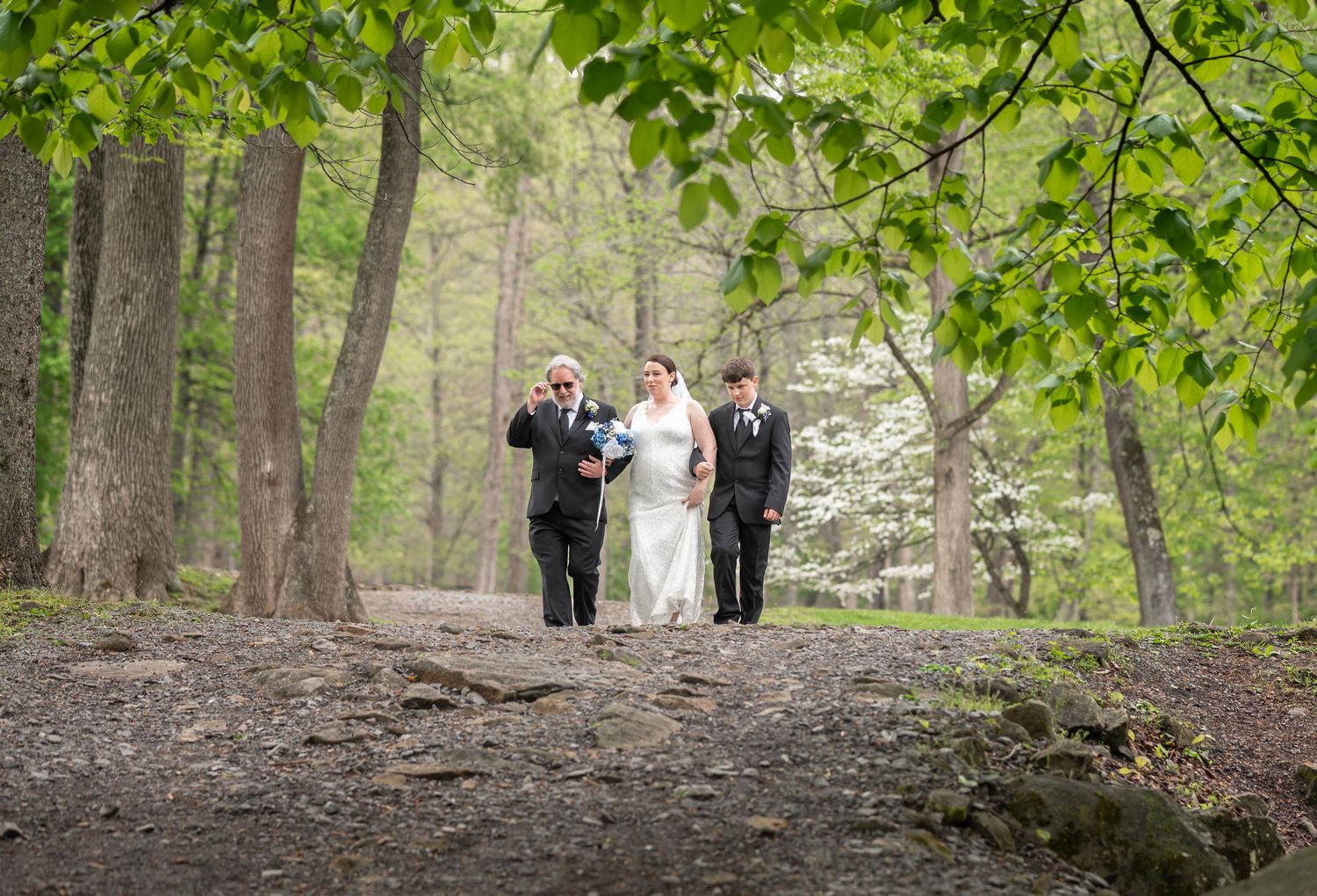 a bride walks down a forest path to her wedding ceremony with her father on one side and her son on the other, and trees in the background