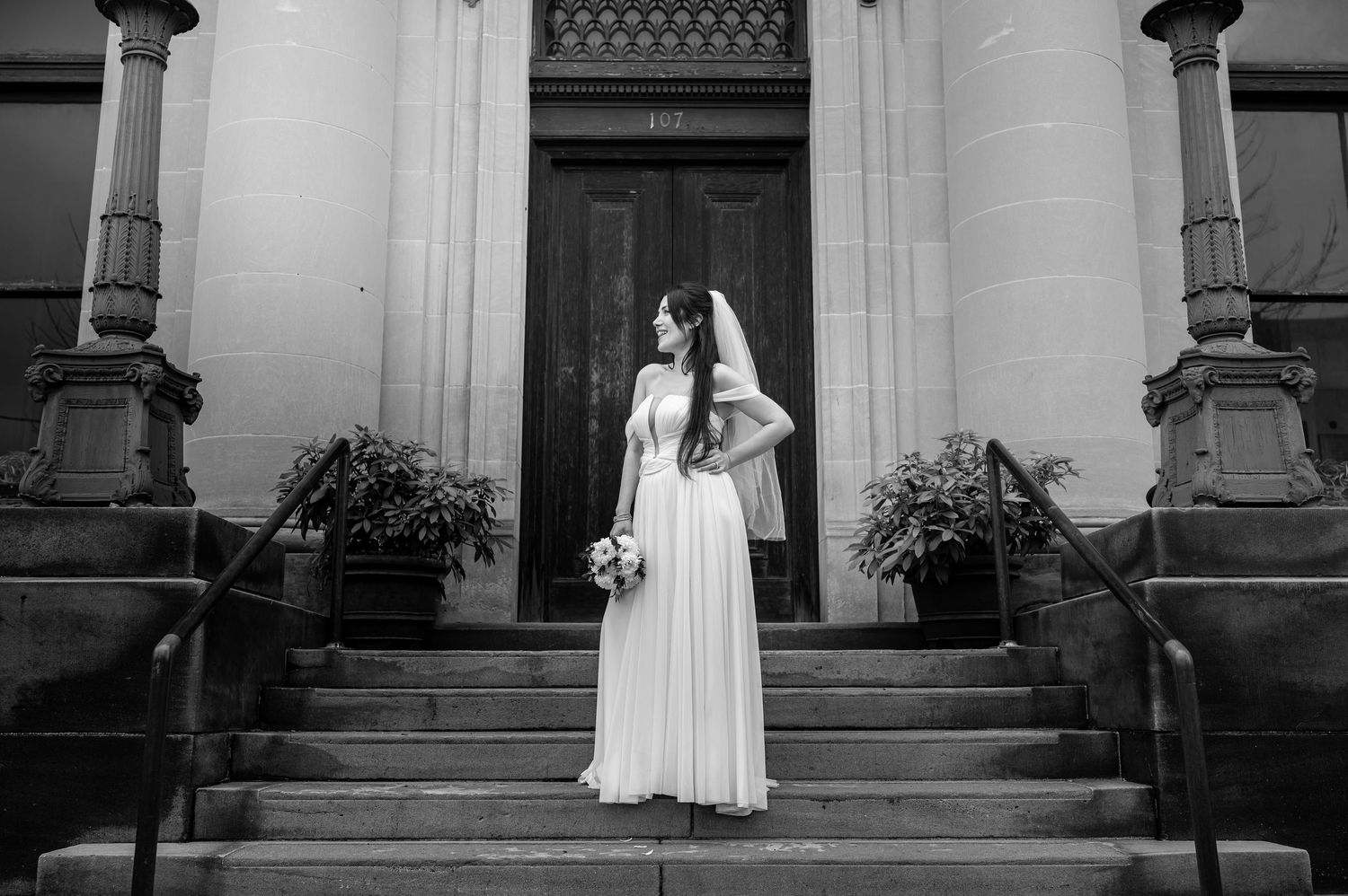 black and white urban portrait of a bride in traditional wedding dress, standing on the stairs of a large building with an old wooden door