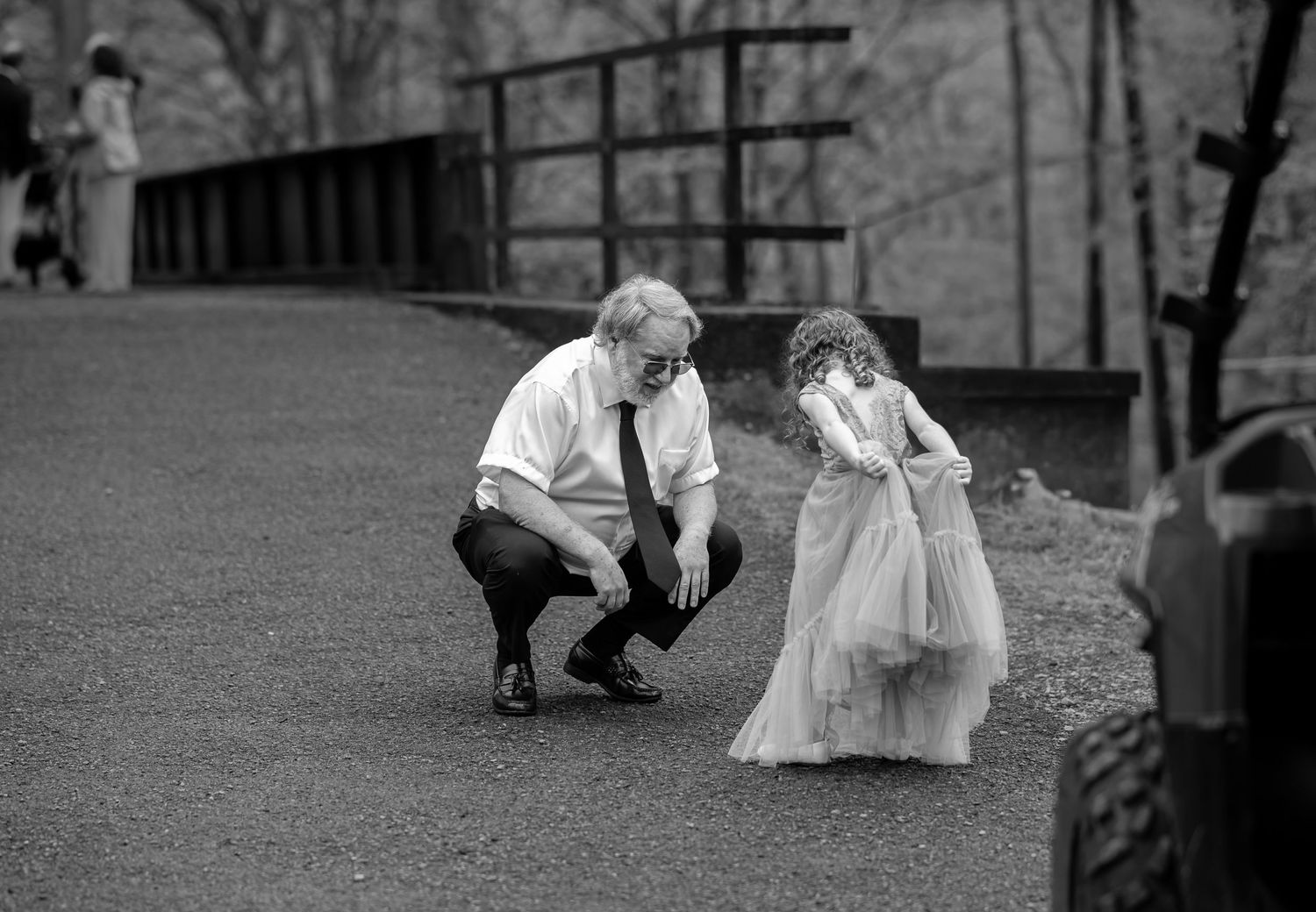 black and white candid image of a grandfather stooping to admire his little granddaughter's fancy flower girl dress, at an outdoor wedding