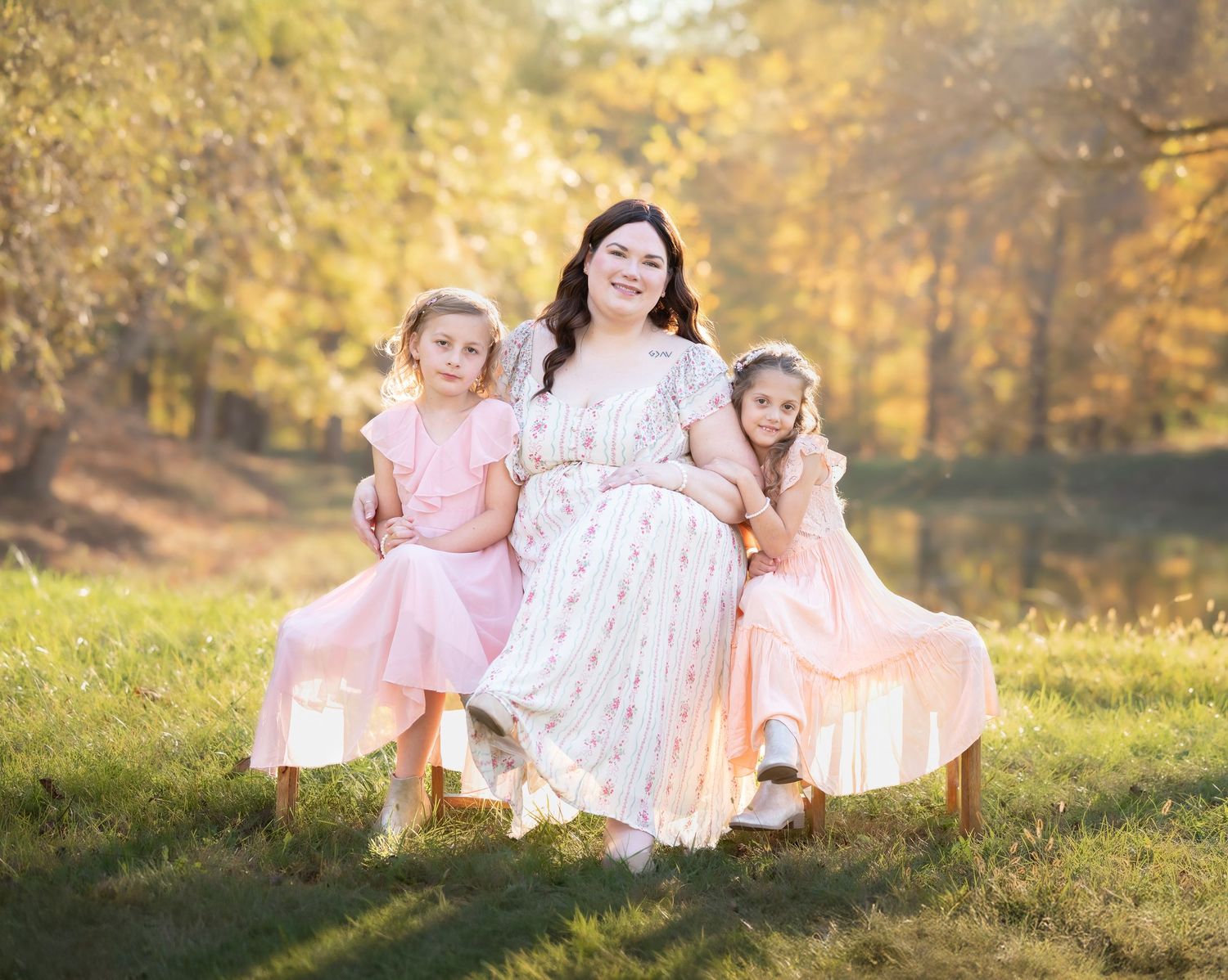 a dark haired woman and her two young daughters, all in pink and white dresses, sit on a bench with glowing gold foliage all around them