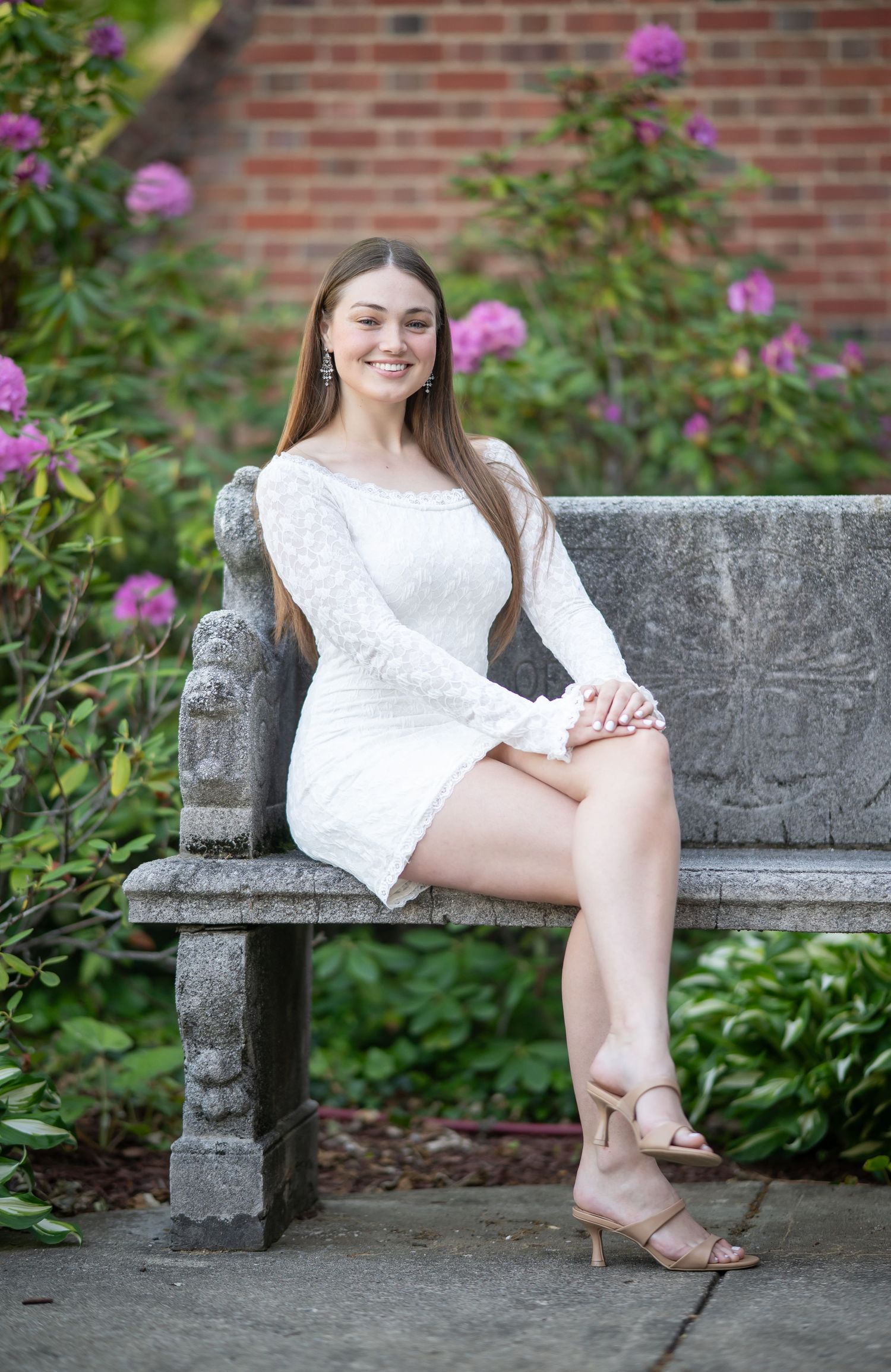 a young WVU graduate in a white dress poses for a senior portrait on a gray stone bench surrounded by flowers on WVU's campus