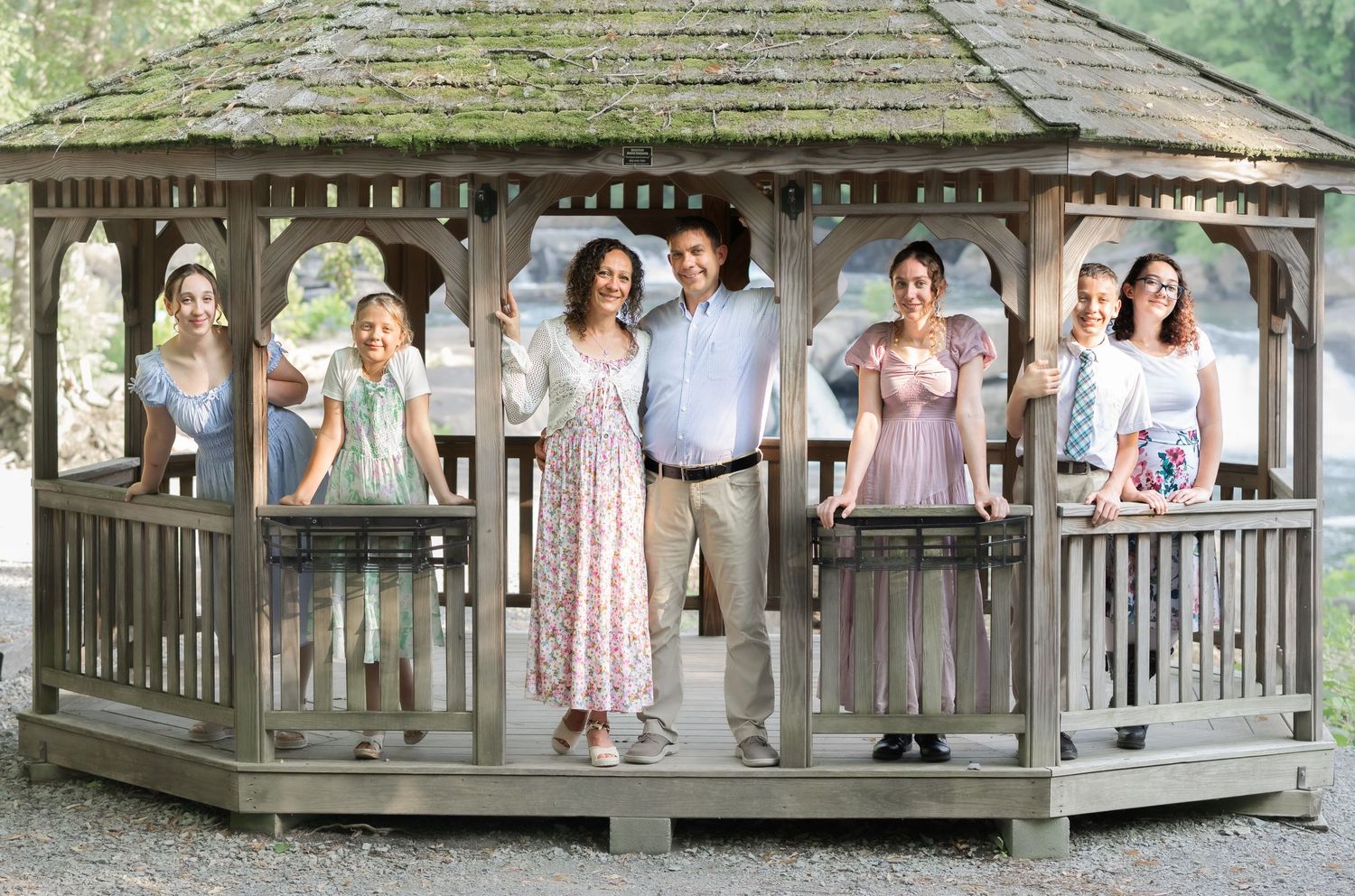 a family of seven poses in various positions in an old wooden gazebo in front of a waterfall, and smiles for a portrait, in Valley Falls, WV
