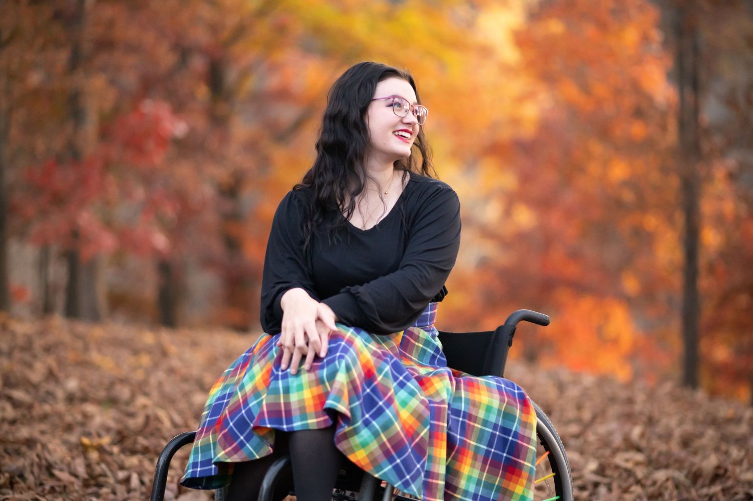 a young woman with dark hair in a black top and colorful plaid skirt poses for a portrait in her wheelchair and laughs