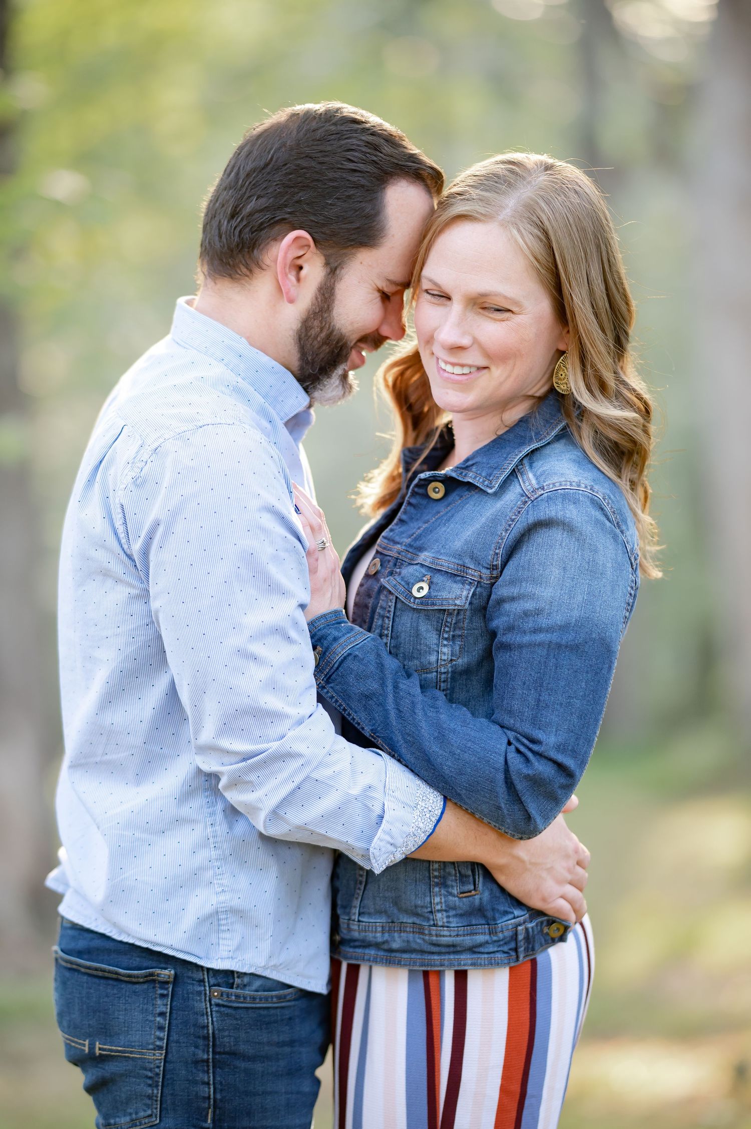 a husband and wife embrace and laugh, with soft sunlit trees and grass in the background; shot during a family session in Morgantown, WV