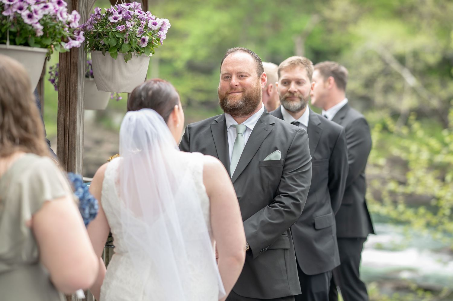 a groom smiles with love at his bride as they stand in front of a gazebo for their wedding ceremony and their bridal party looks on
