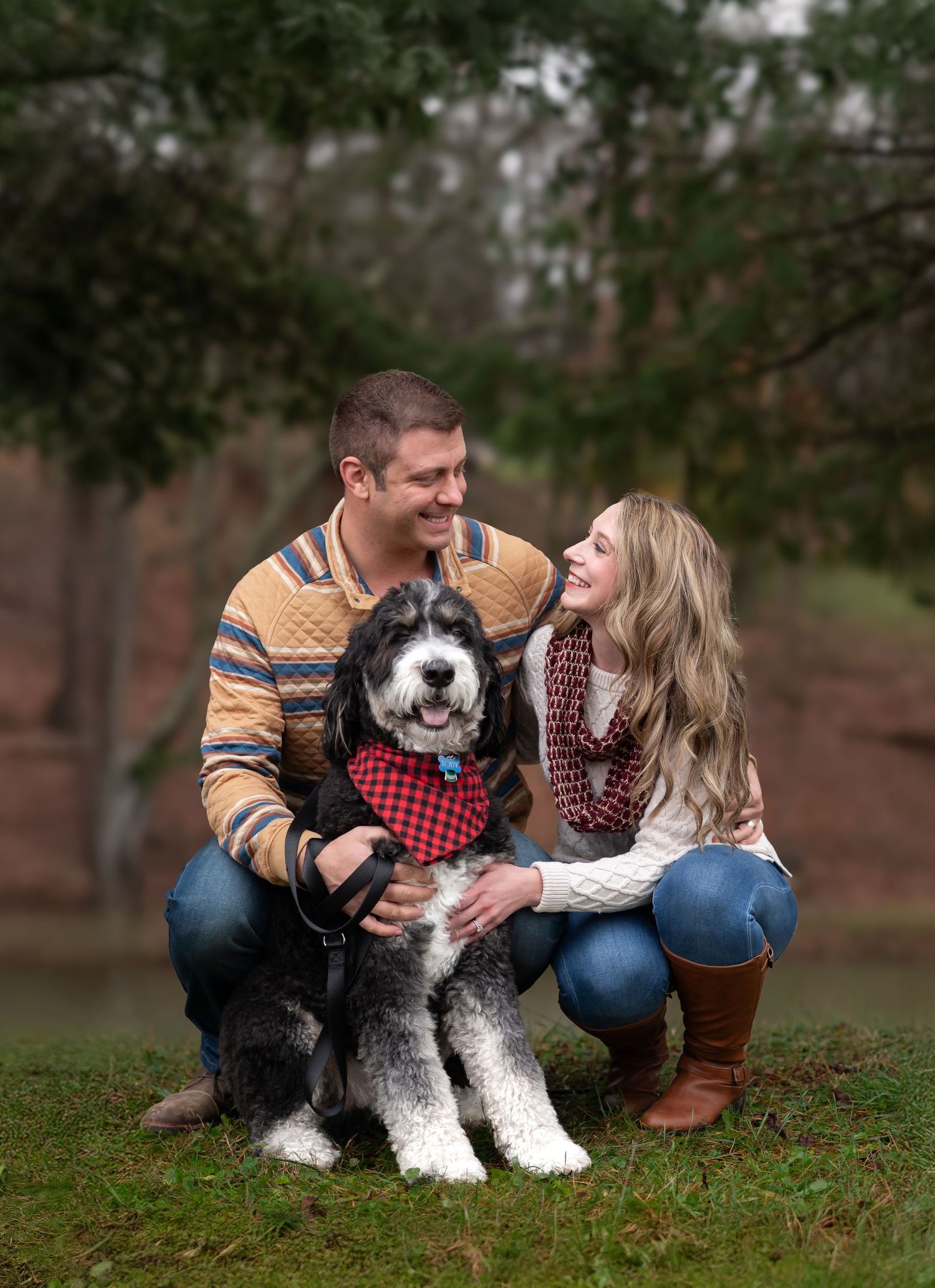 a young couple poses for Christmas card portraits on the ground with their adorable dog in a Christmas scarf; with evergreen trees behind.
