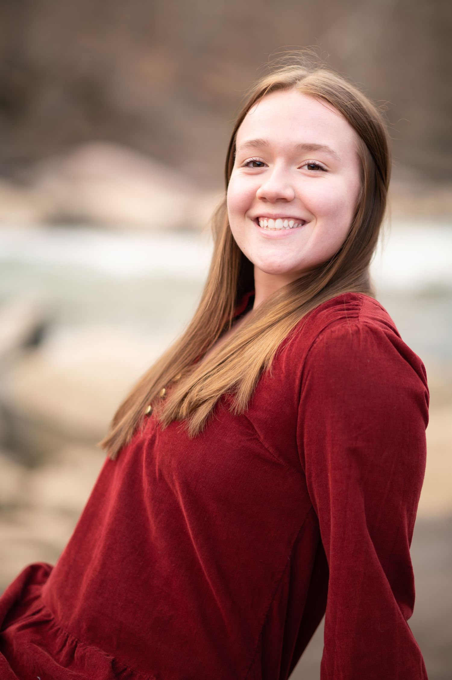 a happy young woman in a red sweater sits in front of a roaring river and smiles into the camera