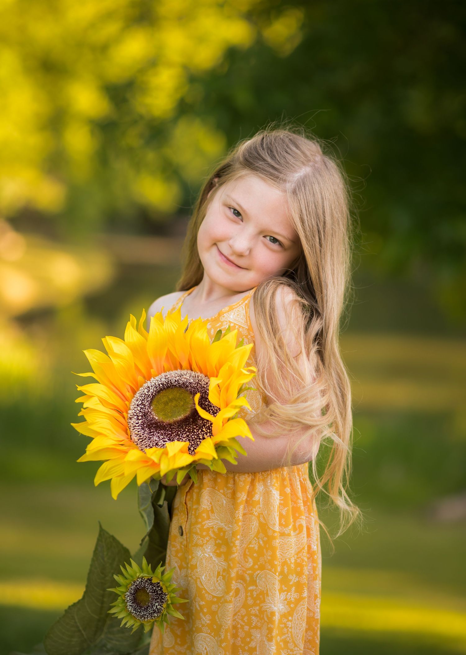 a little girl in a bright yellow dress smiles and holds a large sunflower, with golden and green trees and grass in the background