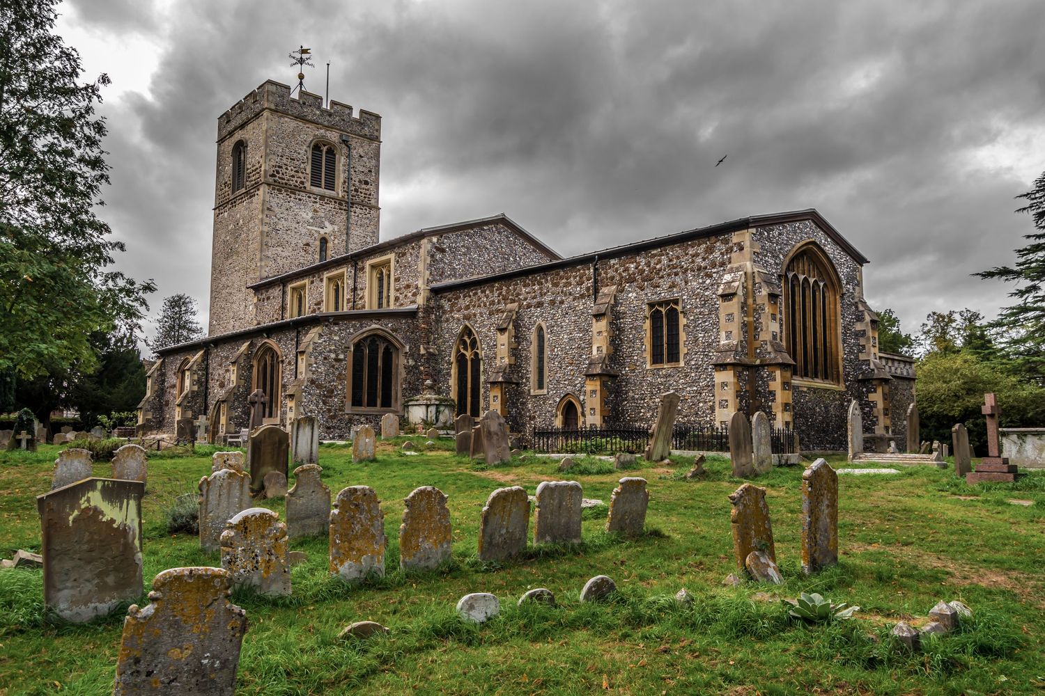 Historic stone church with graveyard featuring weathered tombstones under dramatic cloudy sky in English countryside.