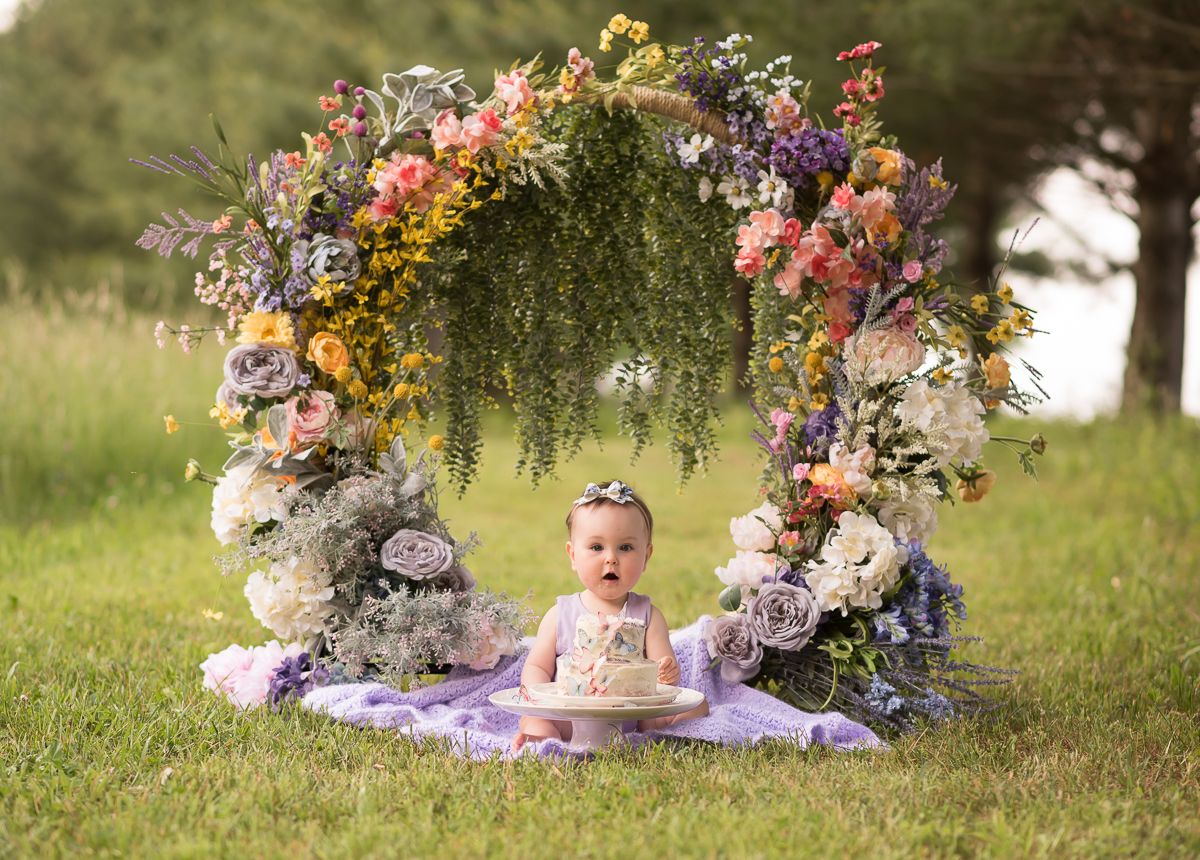 a one year old baby girl in a purple outfit sits on a blanket with her first birthday smash cake under an elaborate arch of spring flowers