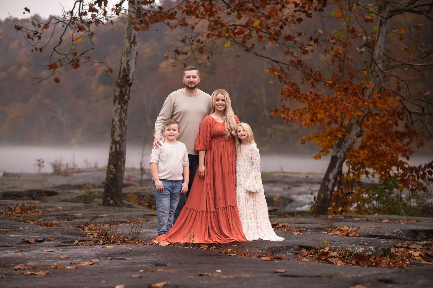 a family of four in coordinating cream and rust outfits poses together for a portrait at the edge of a river with fog and fall leaves behind