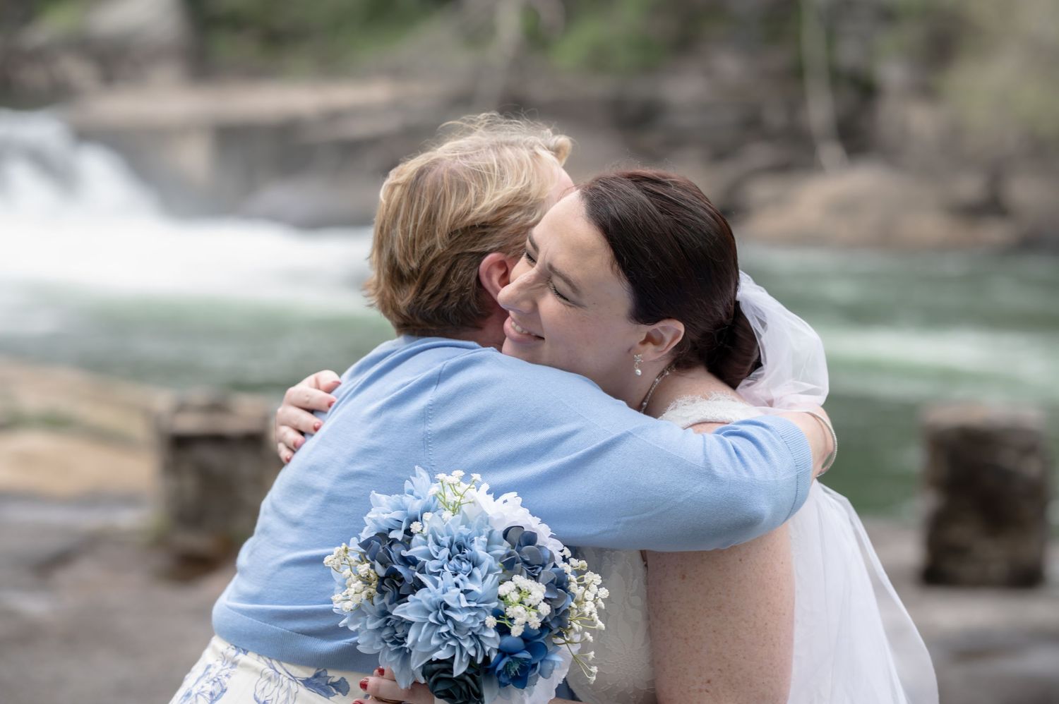 candid shot of a bride in her wedding gown, smiling and embracing the mother of the groom, with waterfalls in the background